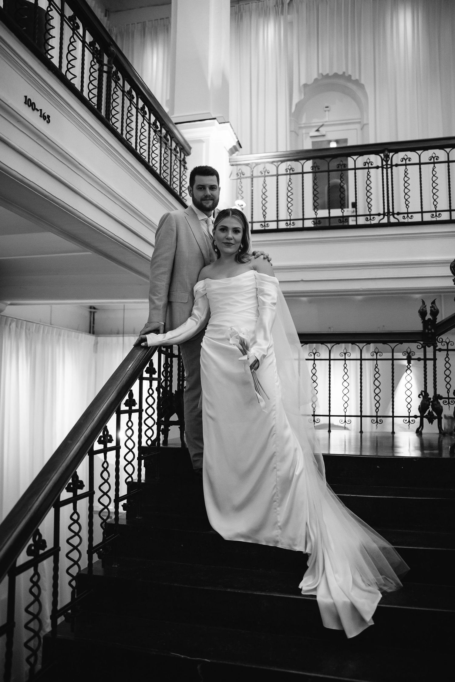 Elegant black and white wedding photo of couple posing on grand staircase with ornate railings.