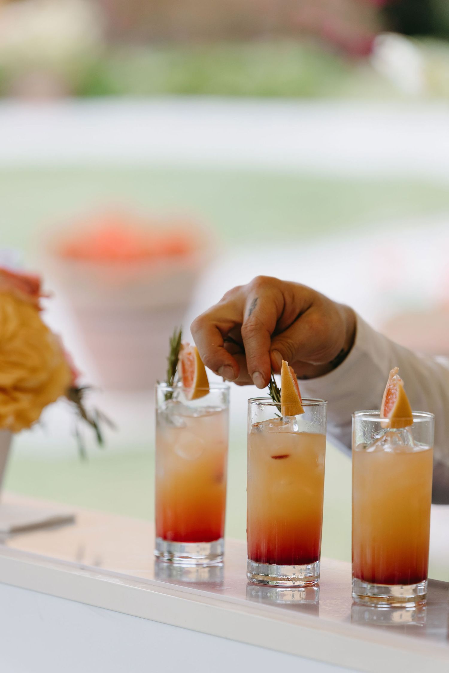 Three layered sunset-colored cocktails being garnished on a white bar.