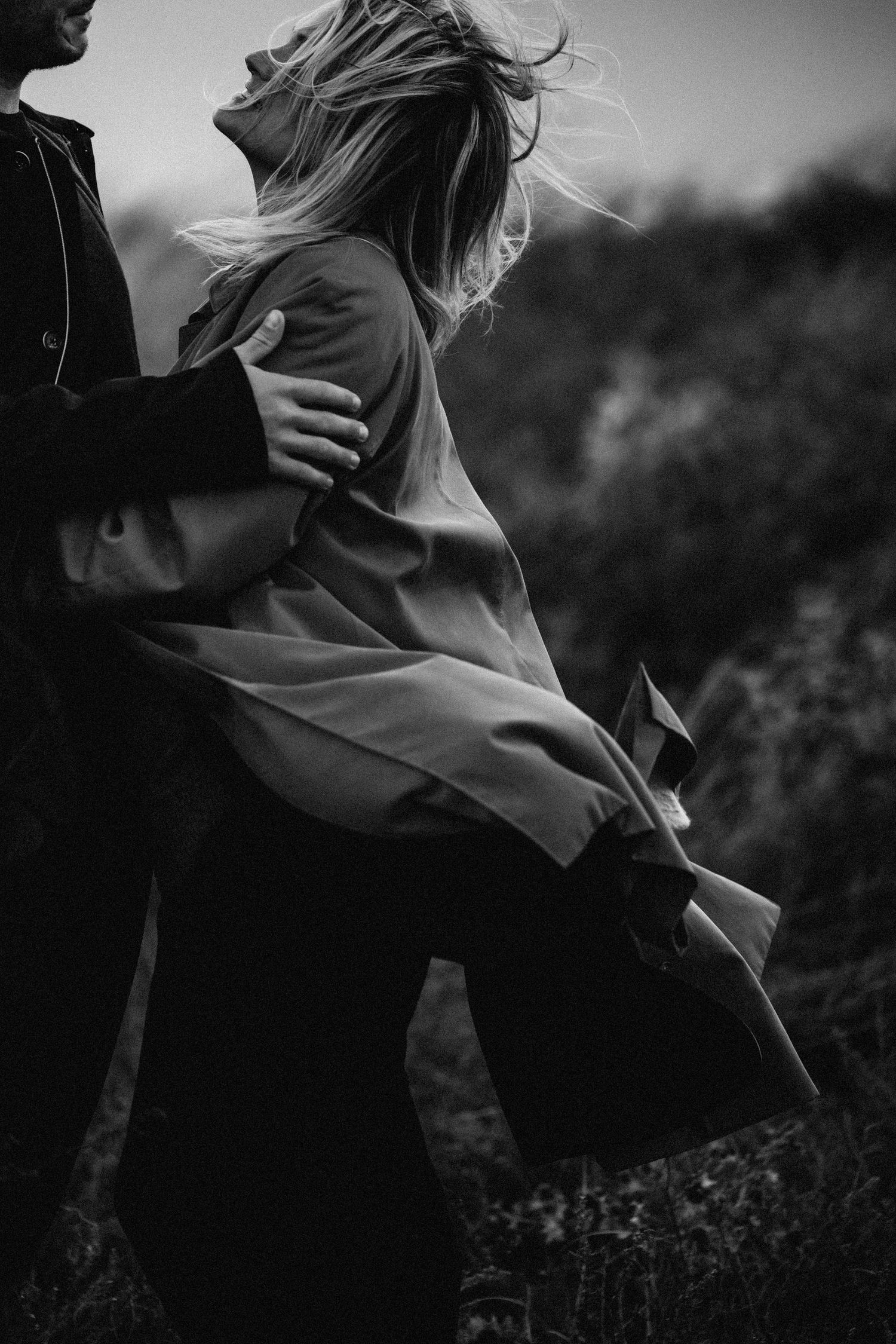 A black and white photo captures a couple's intimate moment in nature with windswept movement.