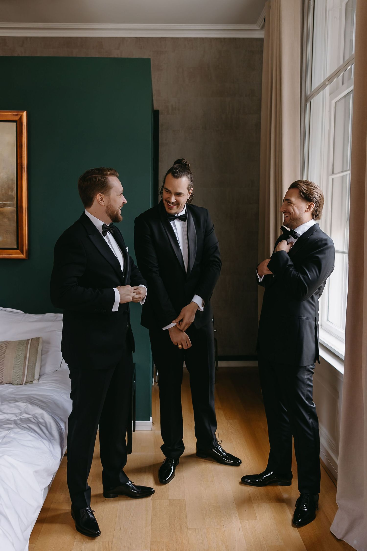 Three men in tuxedos share a lighthearted moment while getting ready in a room with green walls.