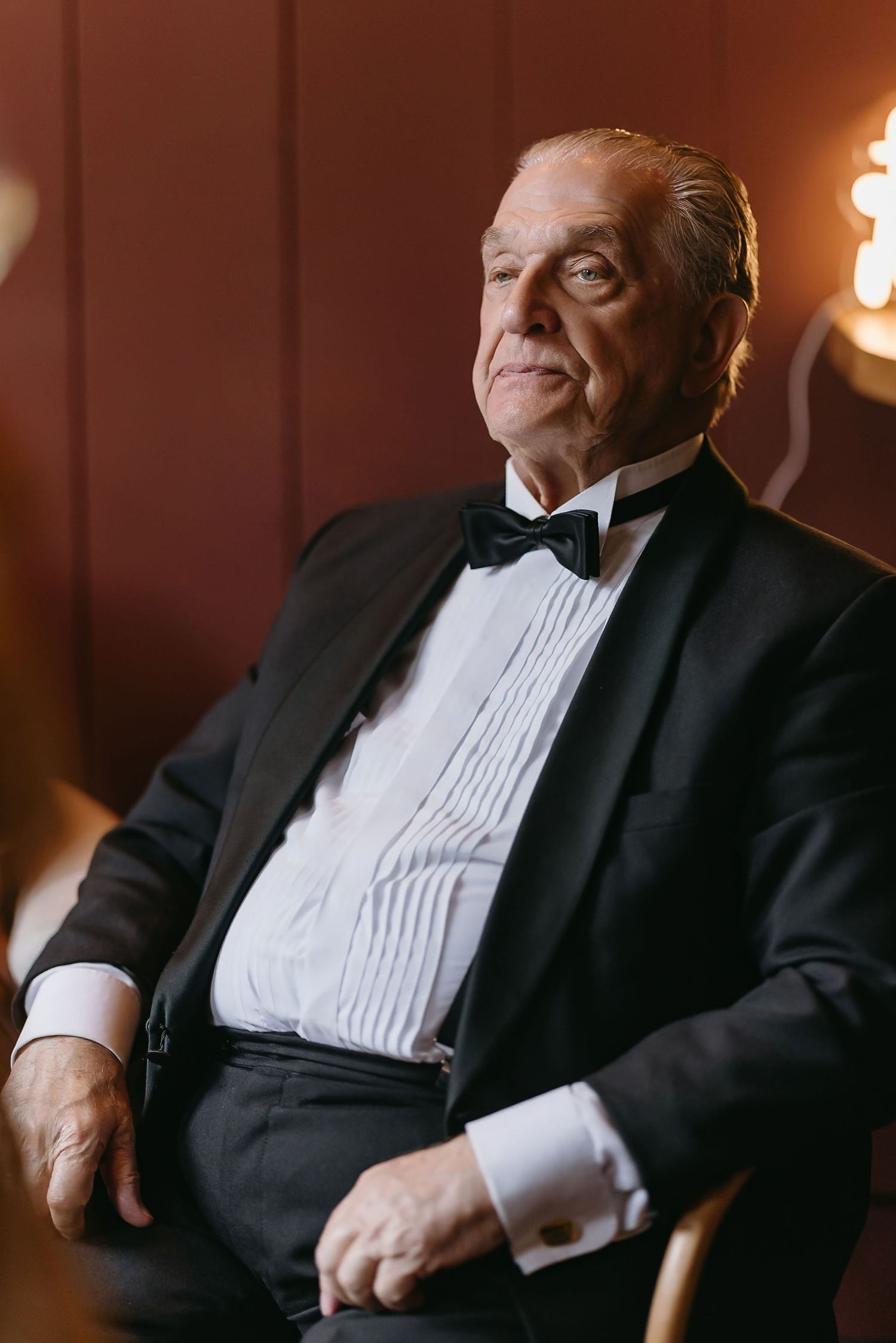 Formal portrait of person in tuxedo and bowtie seated in elegant setting.