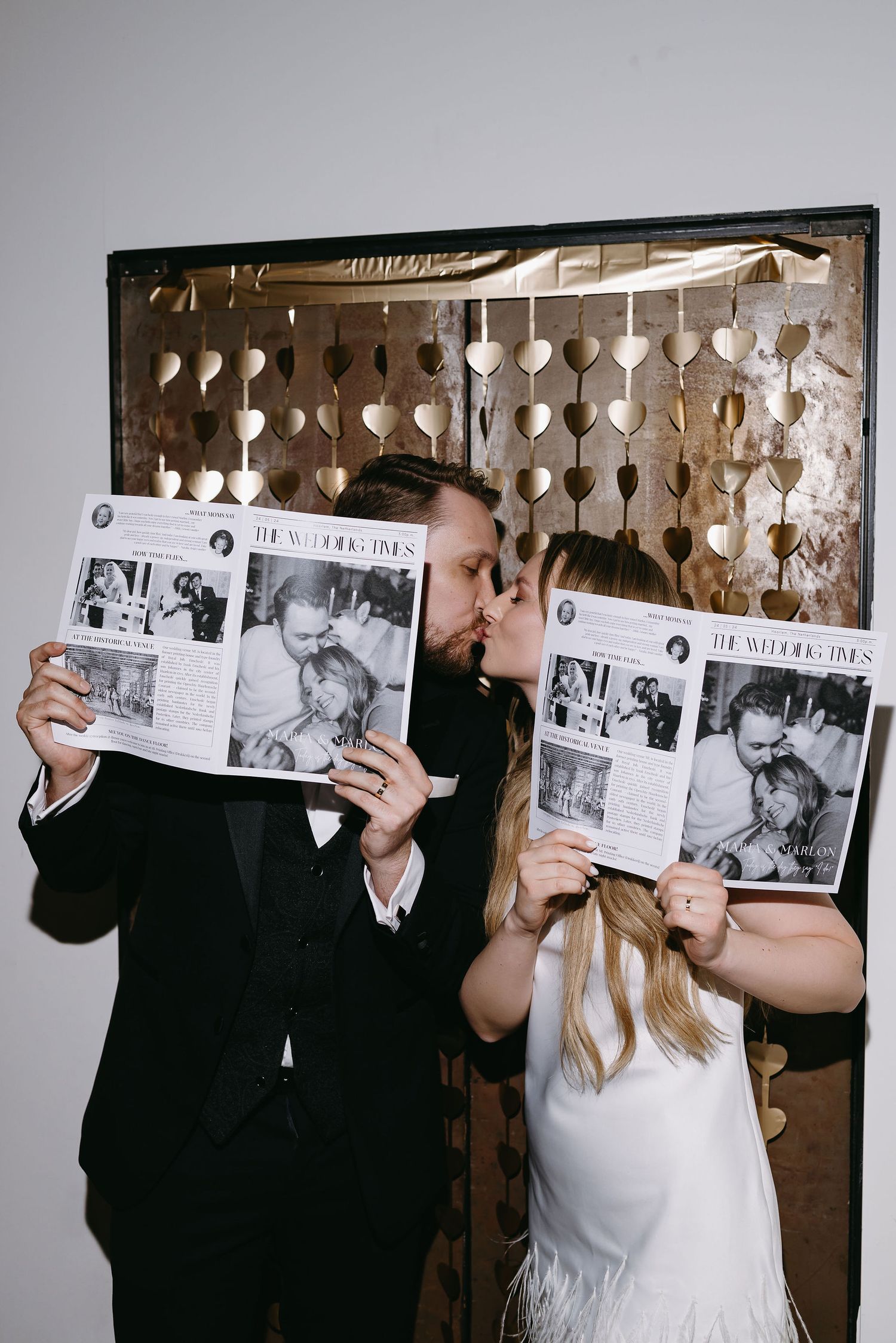 Wedding couple holds up photo prints in front of decorative heart backdrop.