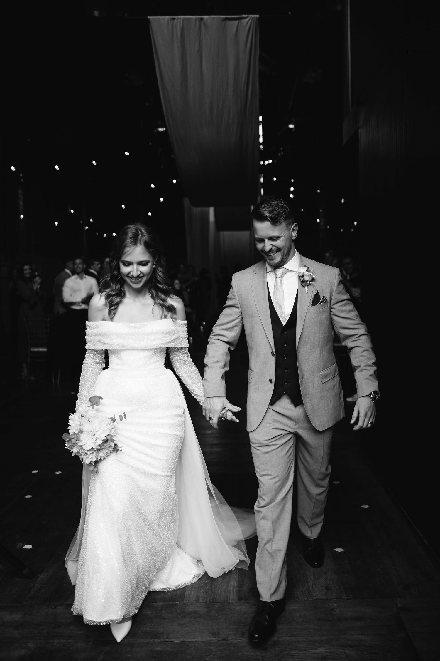 Newlyweds walk hand in hand under twinkling lights in black and white photo.