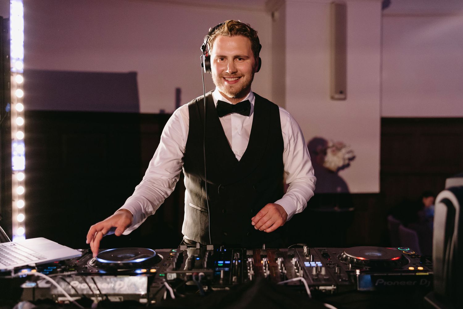 DJ in formal attire mixes music at wedding reception using professional equipment.