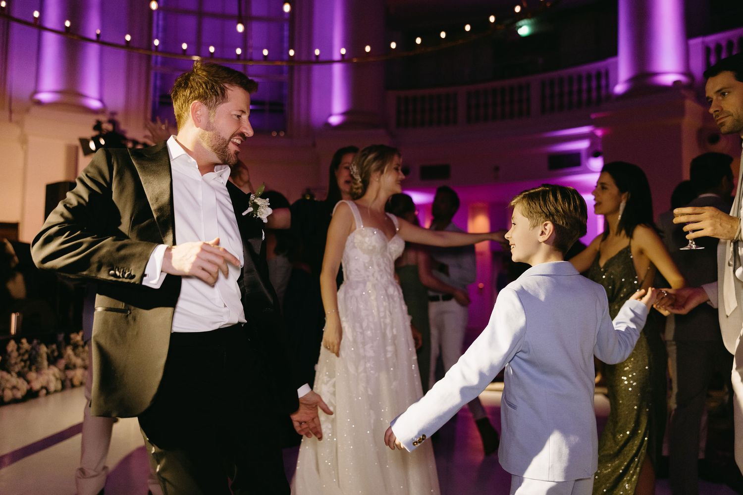 Wedding guests dancing and celebrating on a dance floor illuminated by purple lighting.