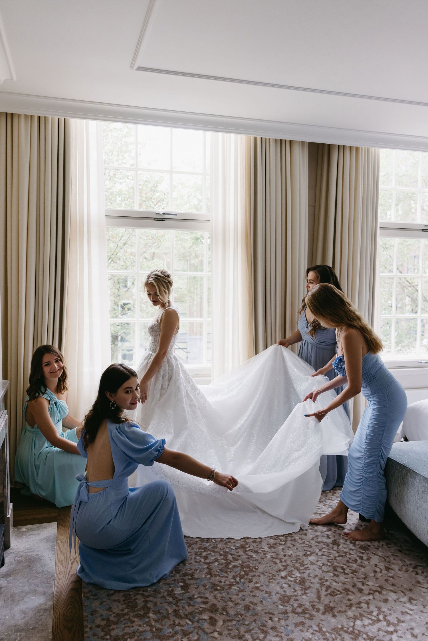 Bridesmaids in blue dresses help arrange wedding gown in sunlit room.