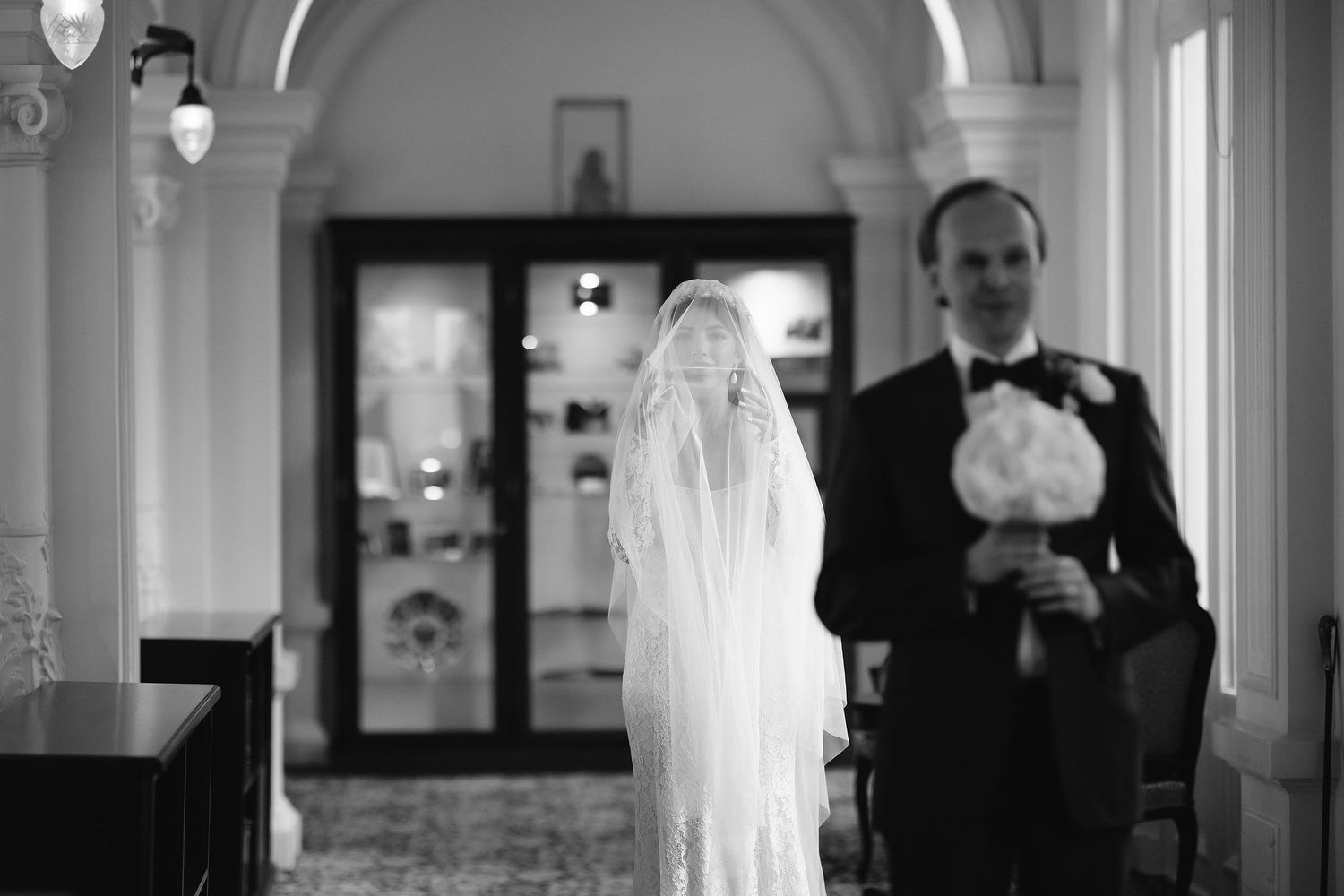 Black and white photo showing couple in wedding attire standing in elegant hallway with arched ceiling and chandeliers.