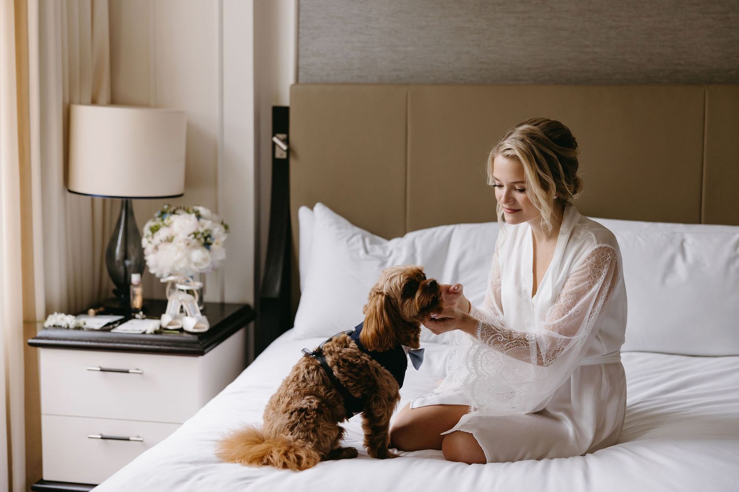 A person in a white robe sits on a hotel bed petting a brown dog while smiling in a bright sunlit room.