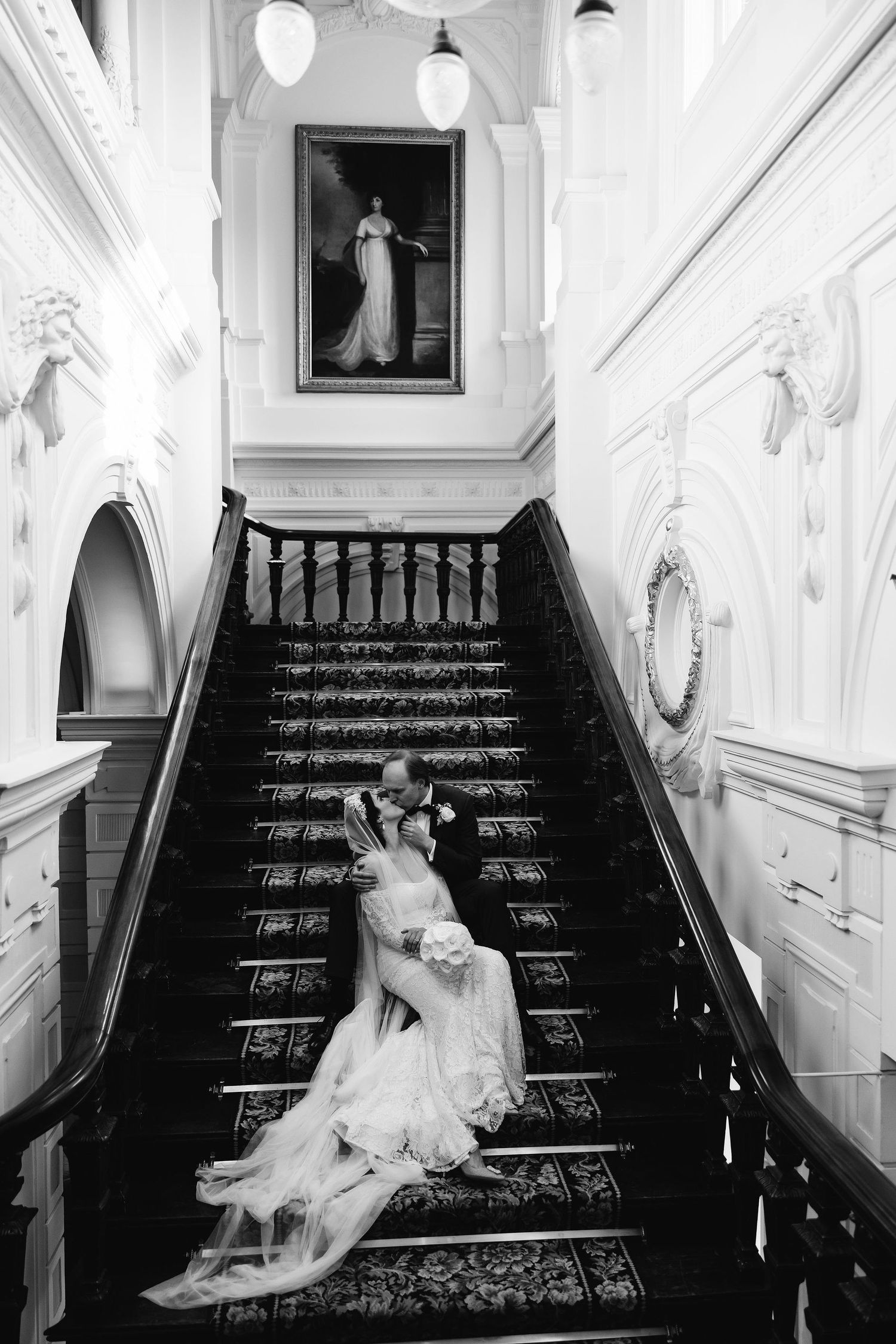 Elegant black and white photograph of a bride reclining on a grand Victorian staircase with ornate carpet runners.