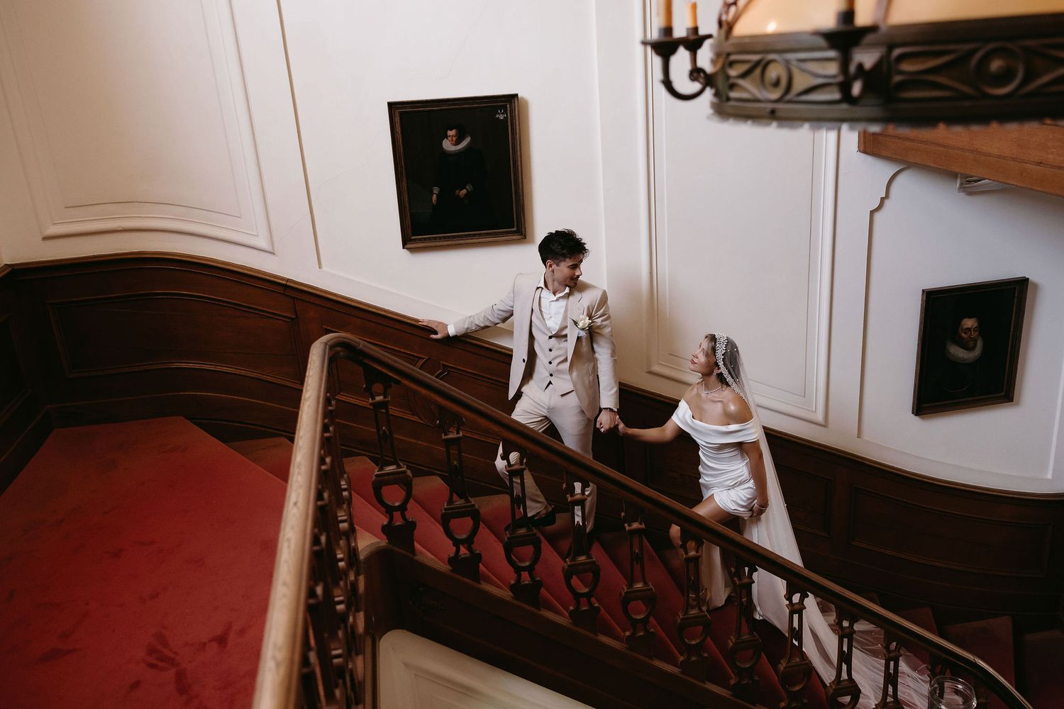 Two people stand on an elegant curved staircase with red carpeting and ornate iron railings in a grand interior space.