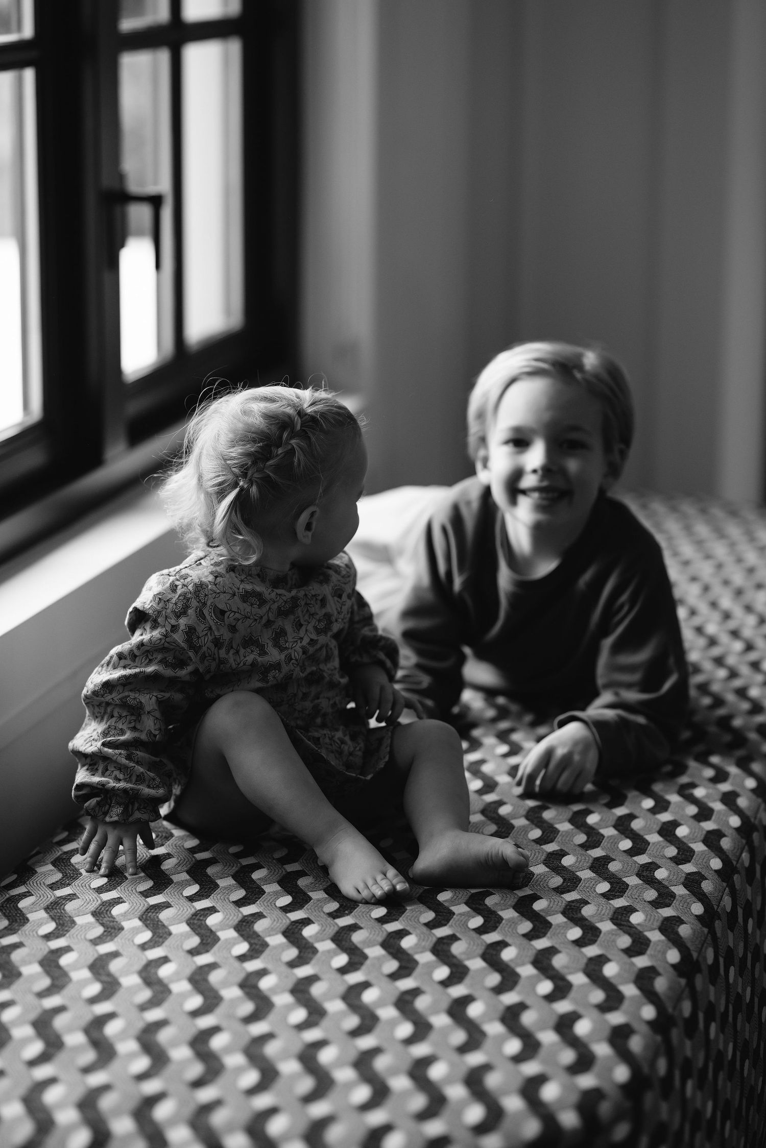 Black and white portrait of children sitting on patterned bedding by window.