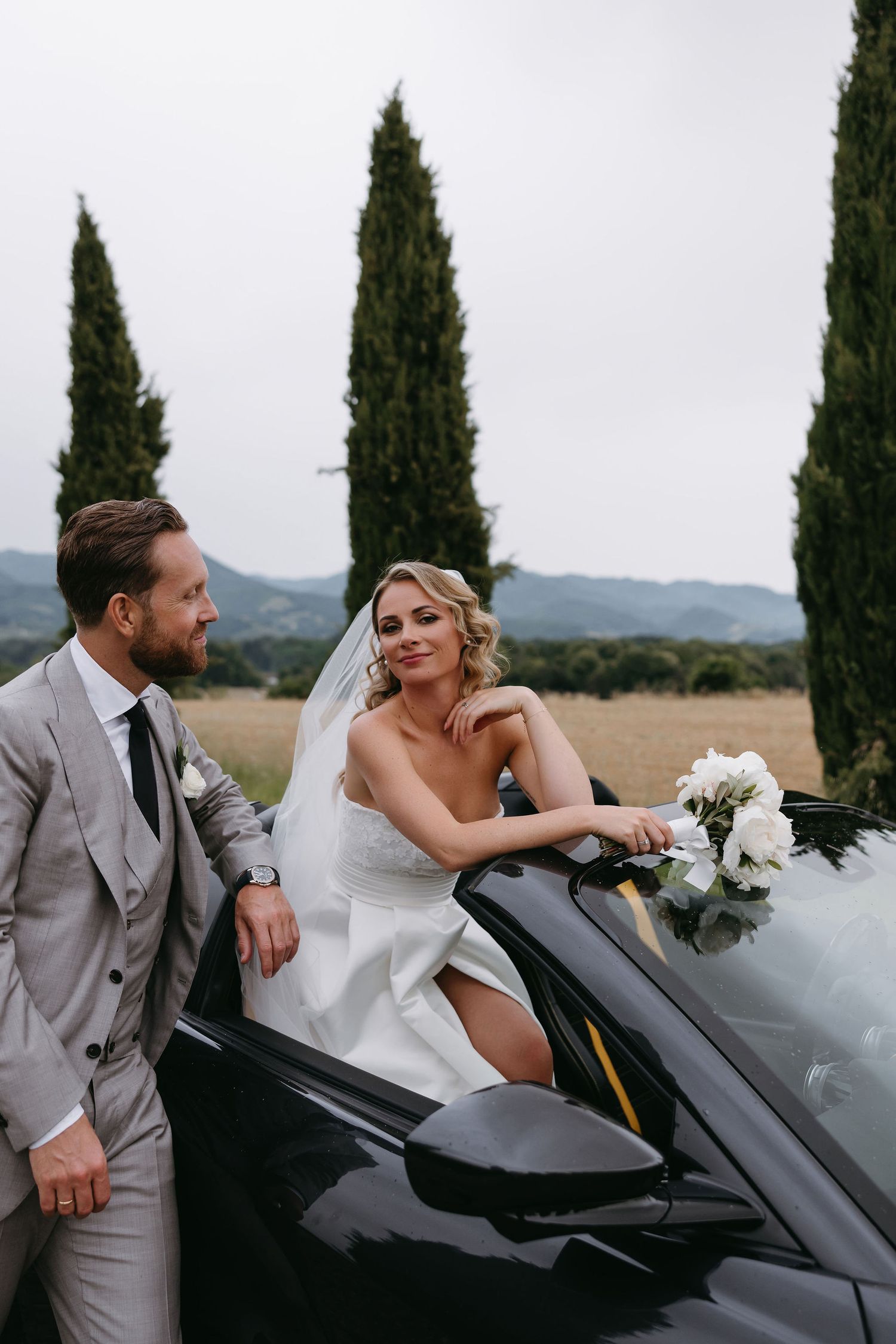 A wedding couple posing with a luxury convertible car against a backdrop of cypress trees.