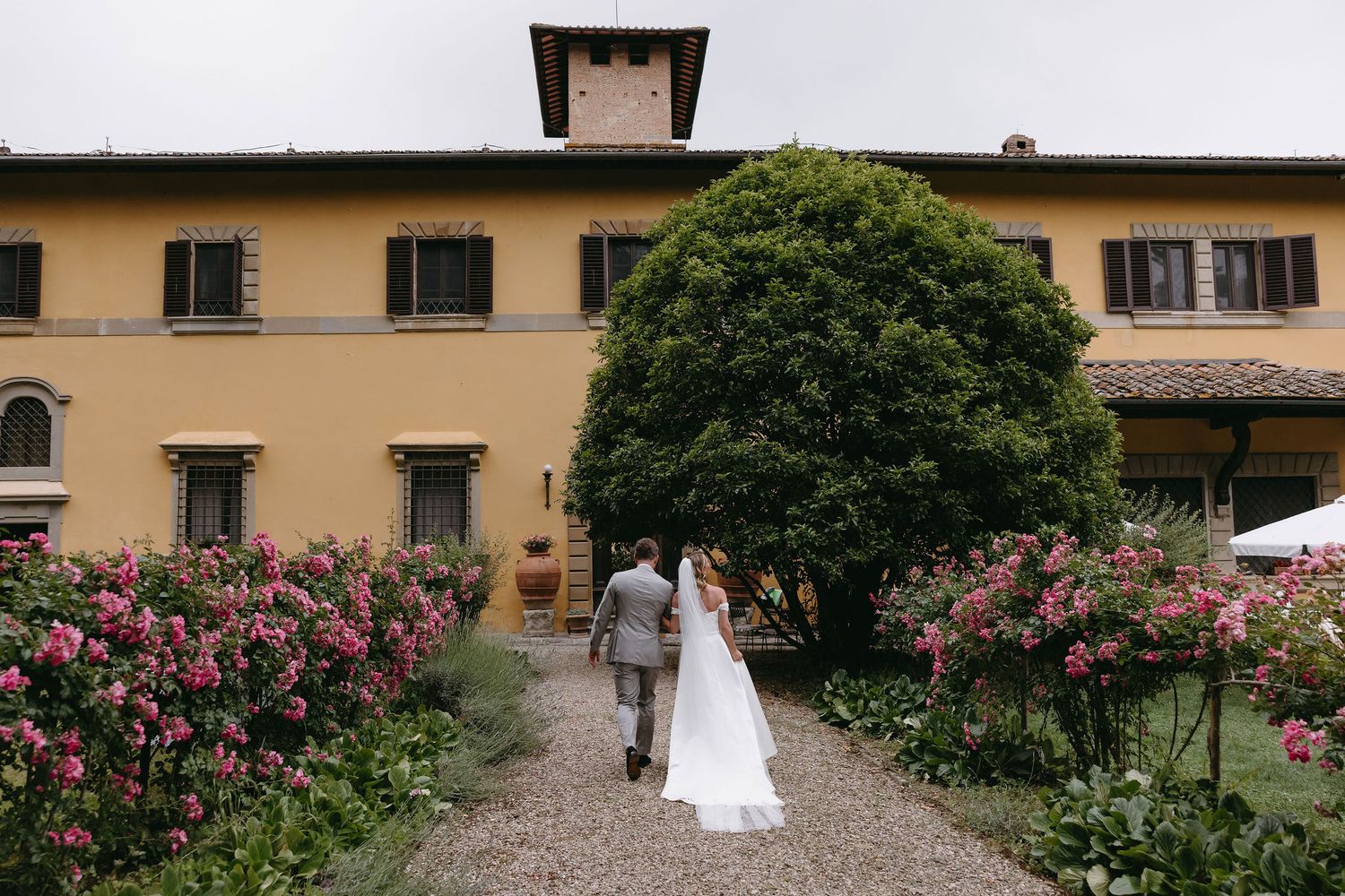 A couple walking down a garden path toward an Italian villa surrounded by blooming flowers.