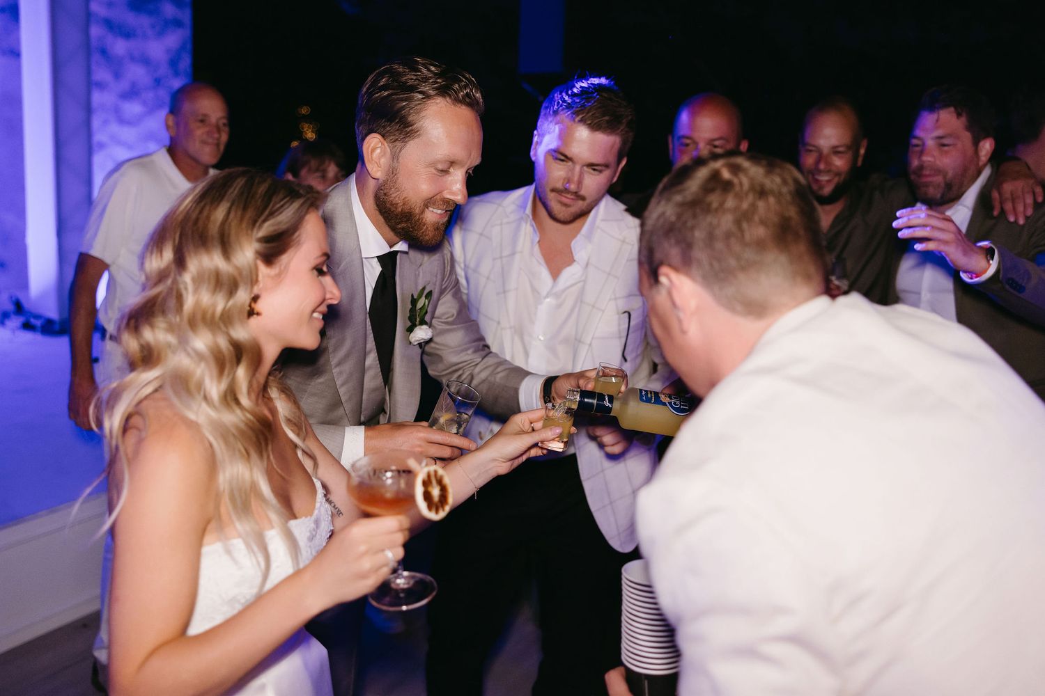 Wedding reception party scene with guests toasting and celebrating under blue lighting.