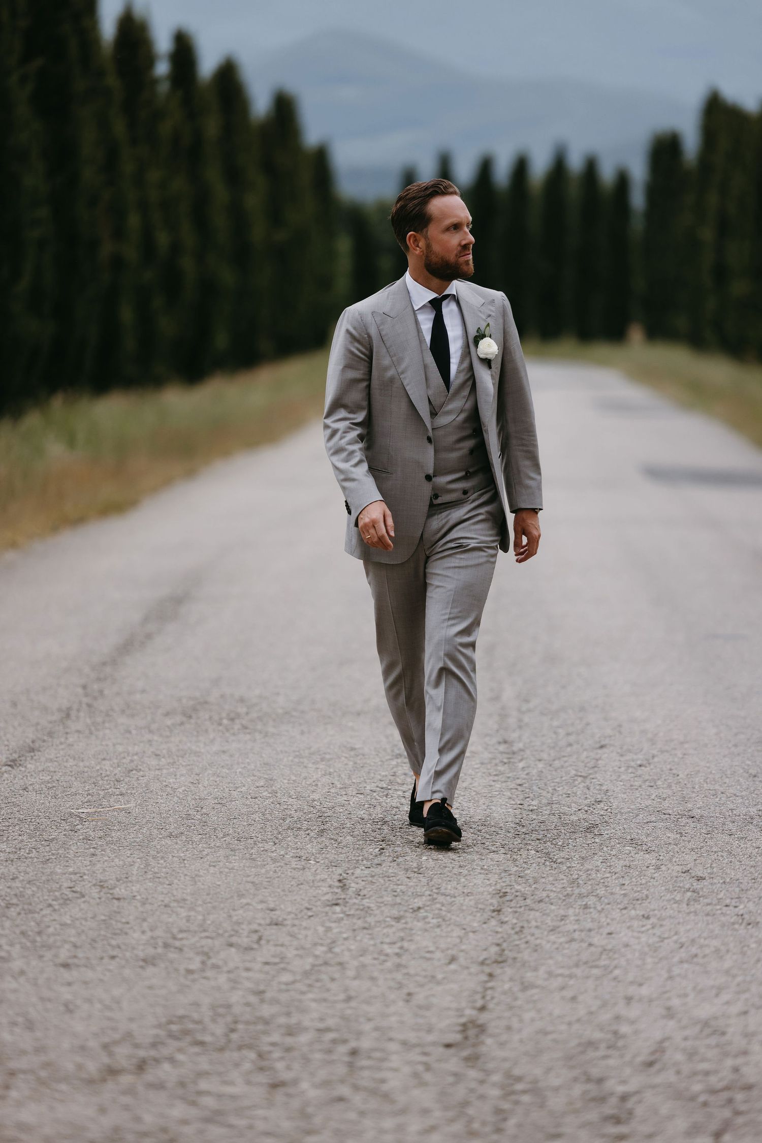 Groom in light gray three-piece suit walking down cypress-lined road in countryside.