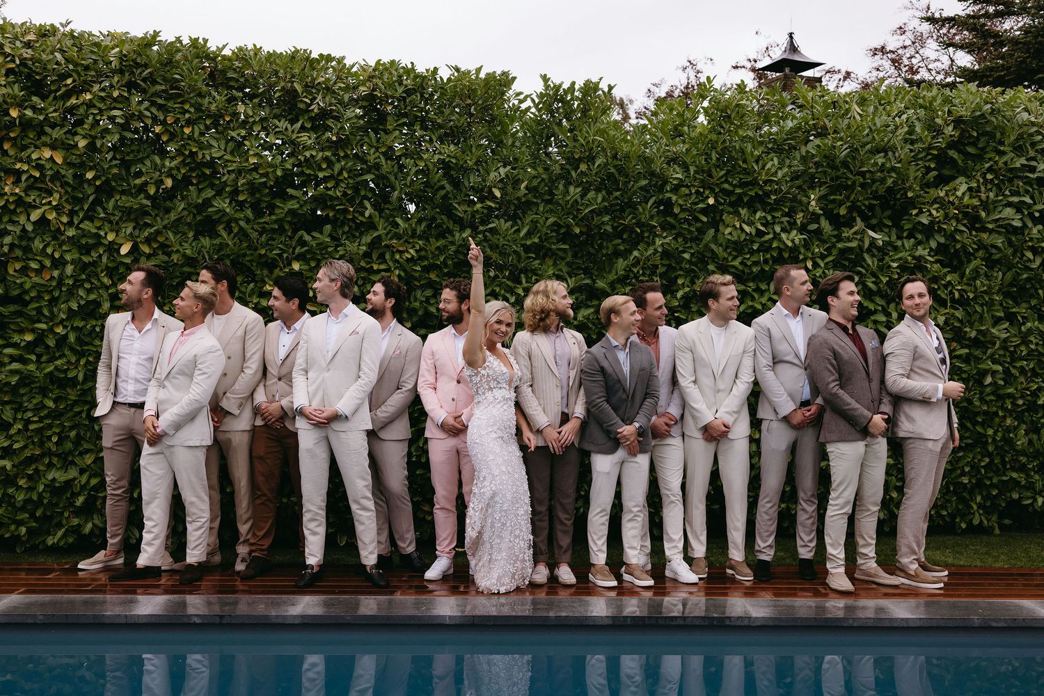 Wedding party in coordinated beige and white attire poses by a pool against a green hedge backdrop.