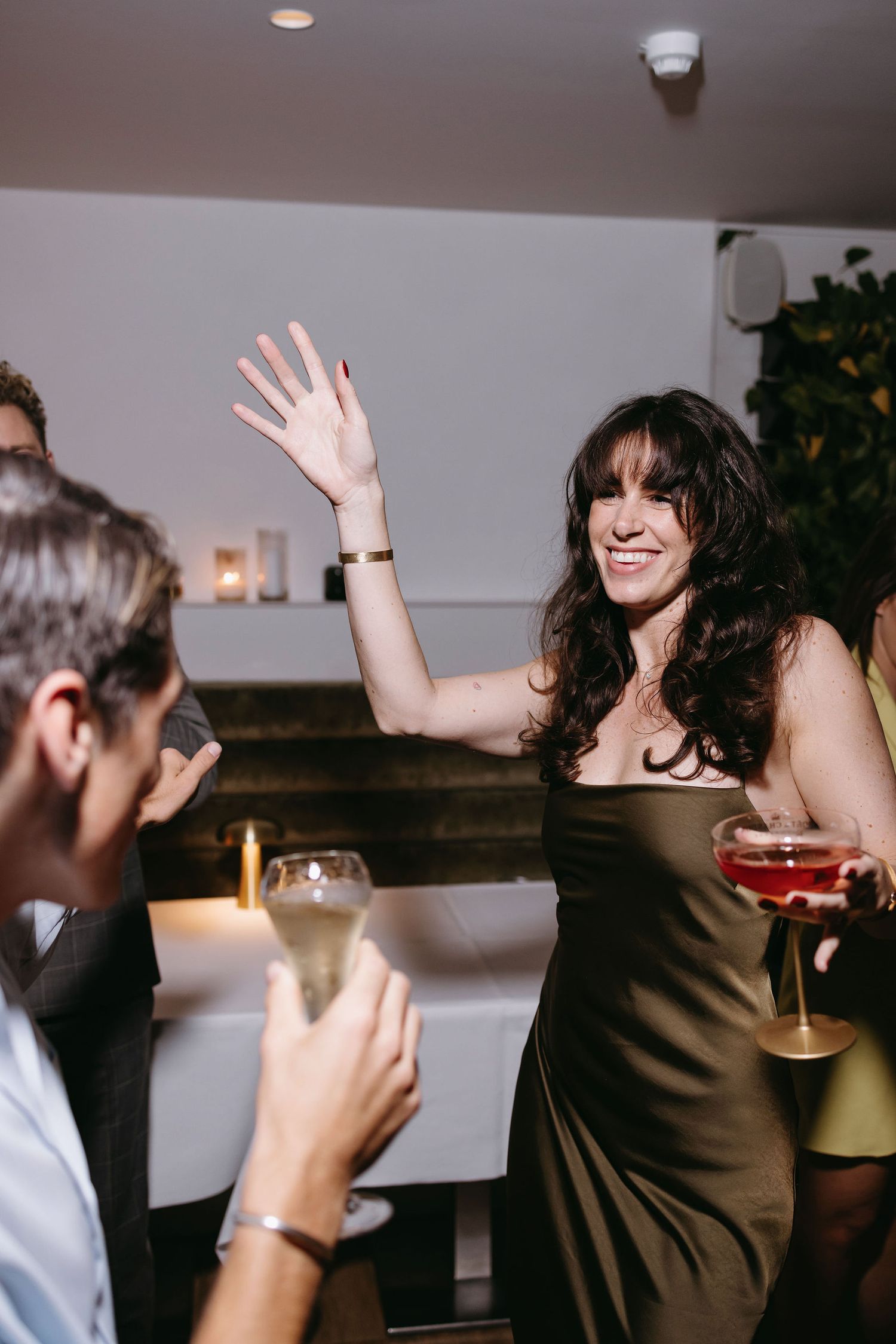 Woman in brown dress raises her hand while socializing at an evening party.