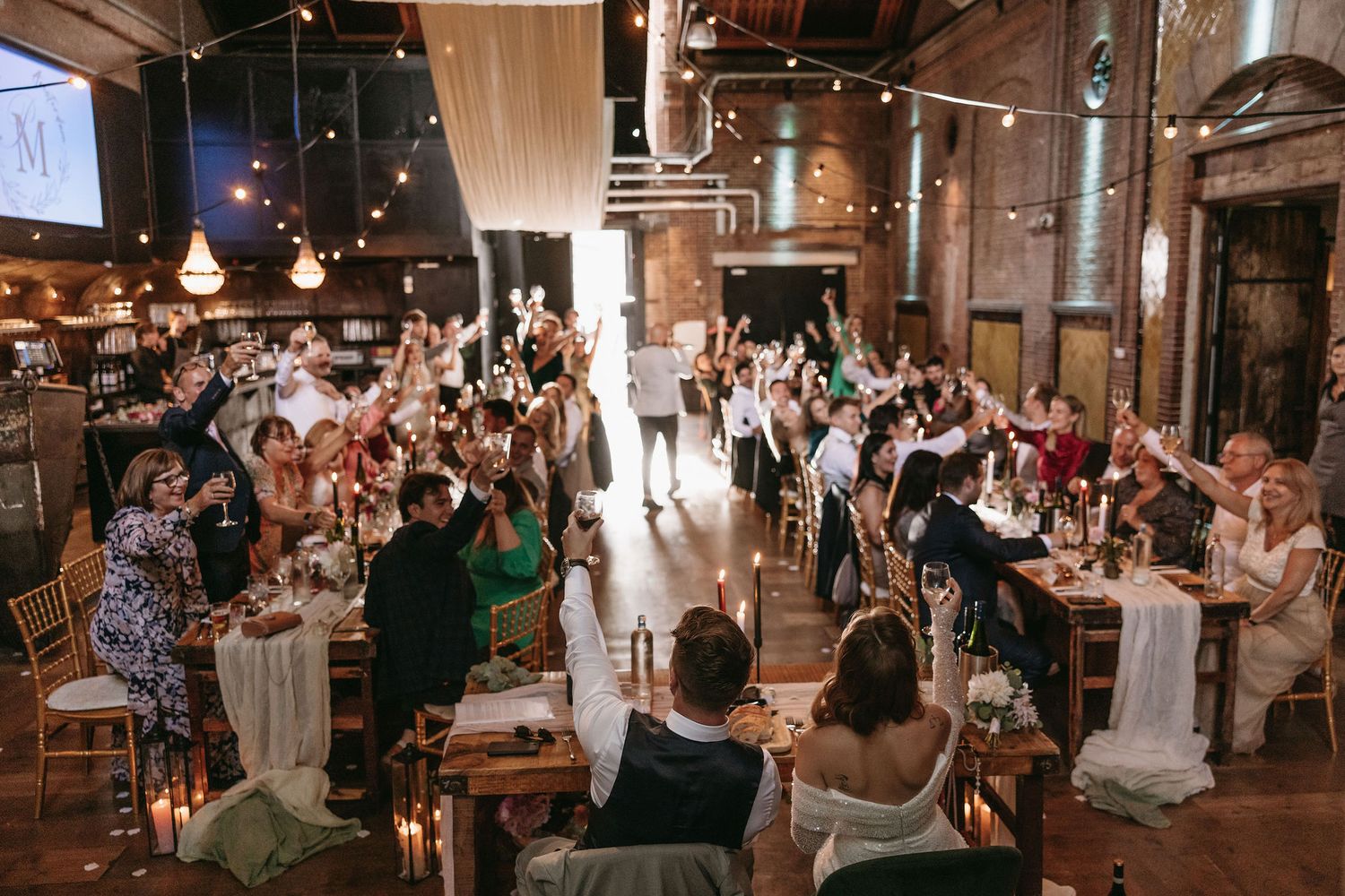 Wedding reception venue filled with guests seated at long tables under hanging lights and white drapery.