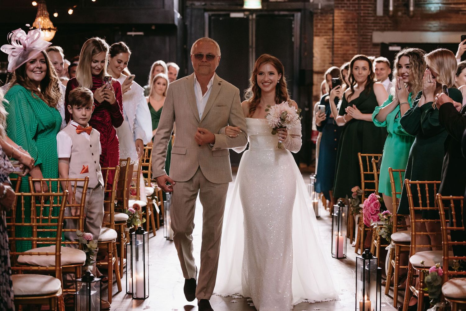 Wedding processional in rustic brick venue with guests seated along aisle.