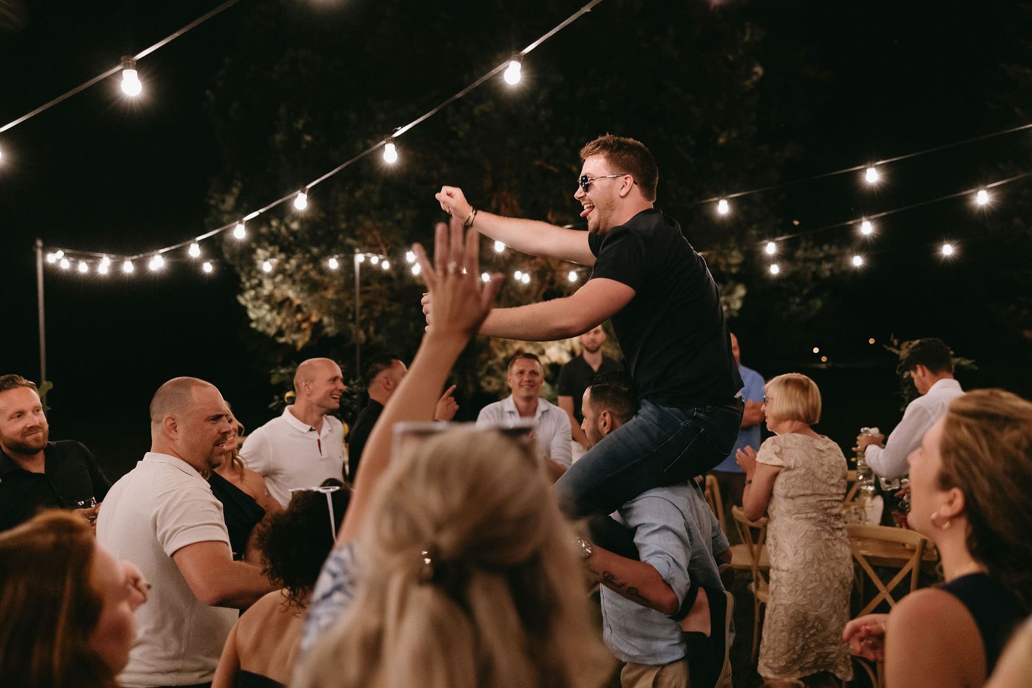 Lively wedding reception celebration with guests dancing under string lights at night.