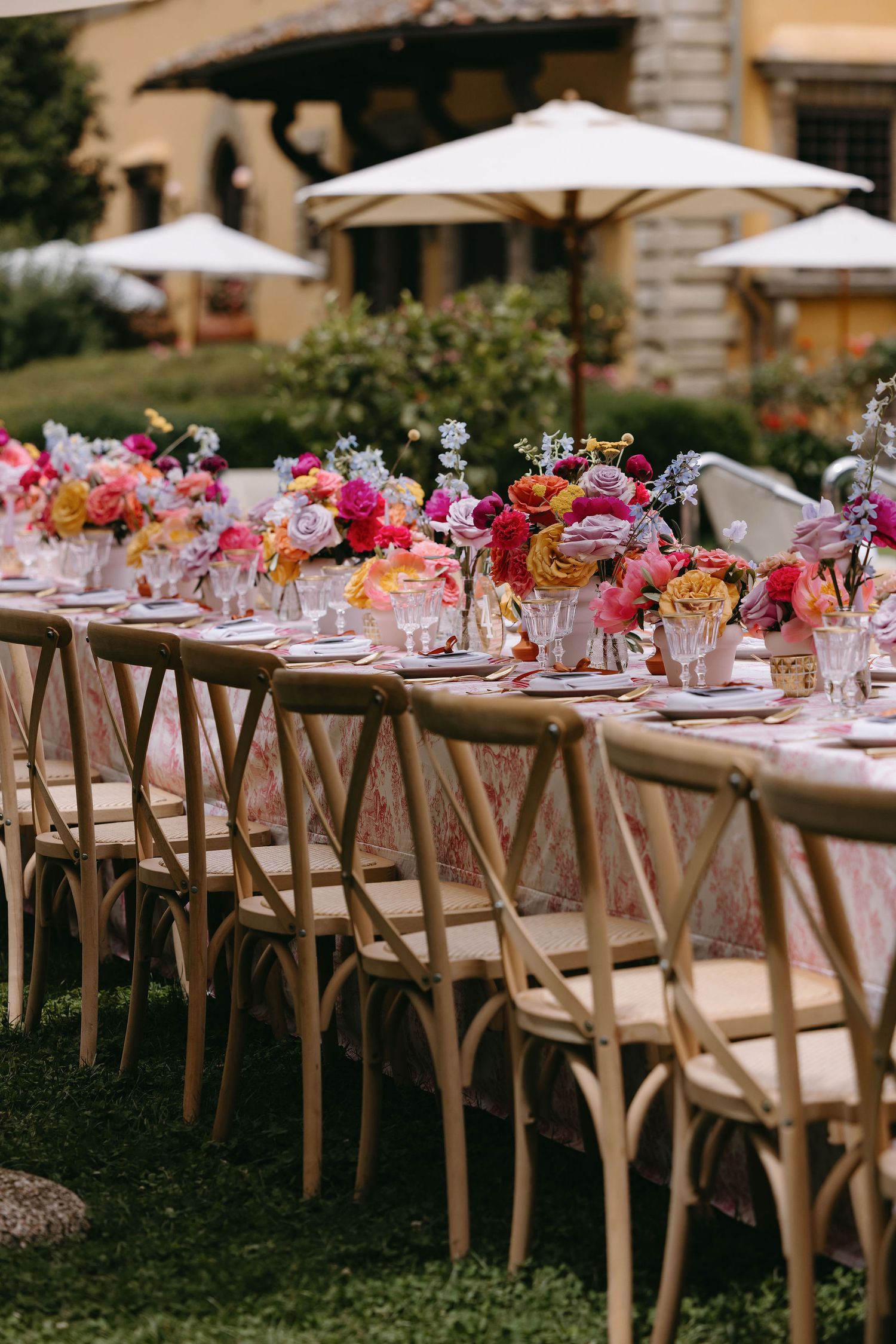 Long outdoor dining table with pink floral arrangements and crossback chairs under white umbrellas.