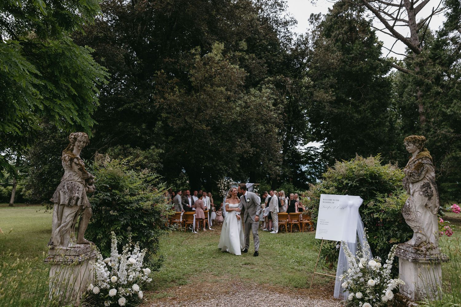 Garden wedding ceremony setup with stone statues and white floral arrangements lining a rustic pathway.