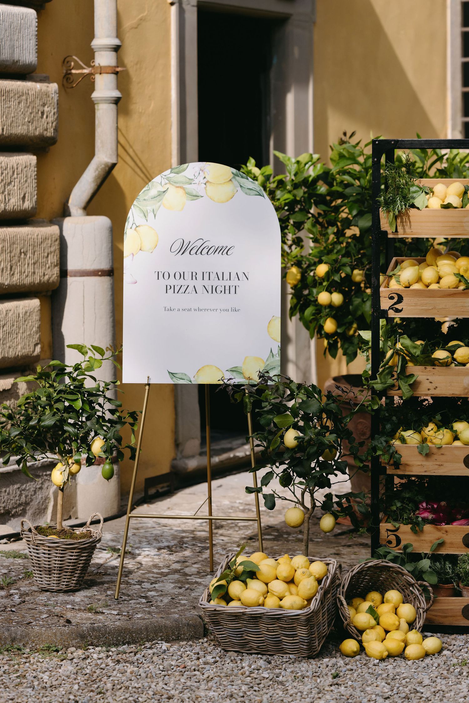 Welcome sign for Italian pizza night surrounded by fresh lemons in wooden crates against a yellow wall.