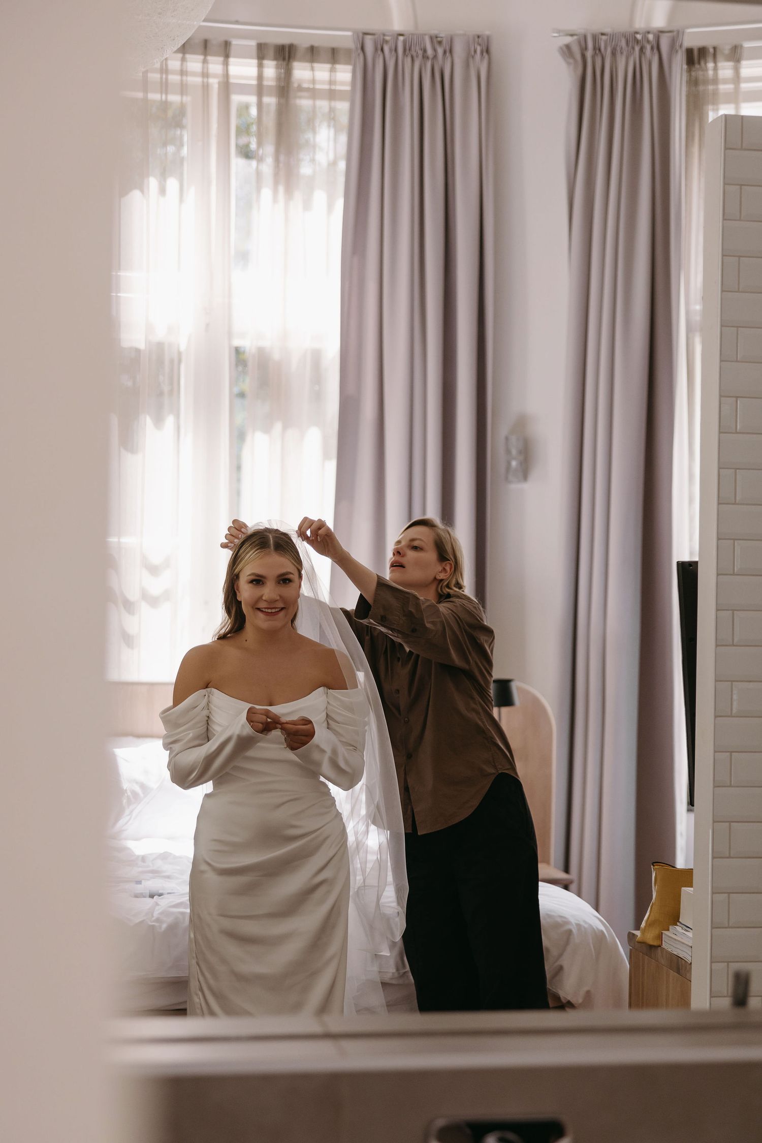 Behind-the-scenes moment of wedding preparation showing final dress adjustments in a sunlit room.