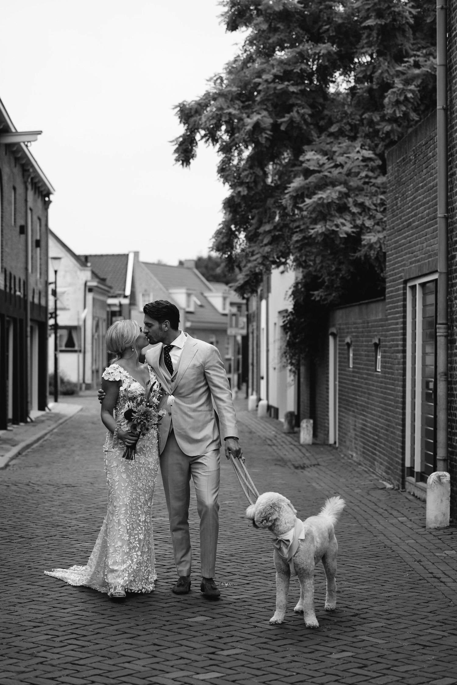 Black and white wedding photos on cobblestone street with couple and white dog.