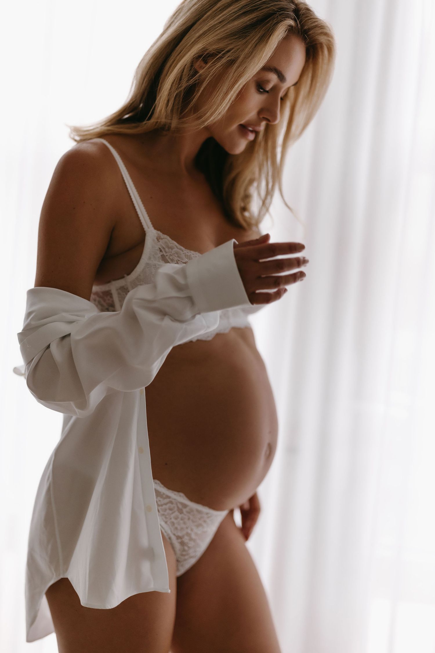 Maternity boudoir photo of expectant mother in white lingerie and open shirt by window.