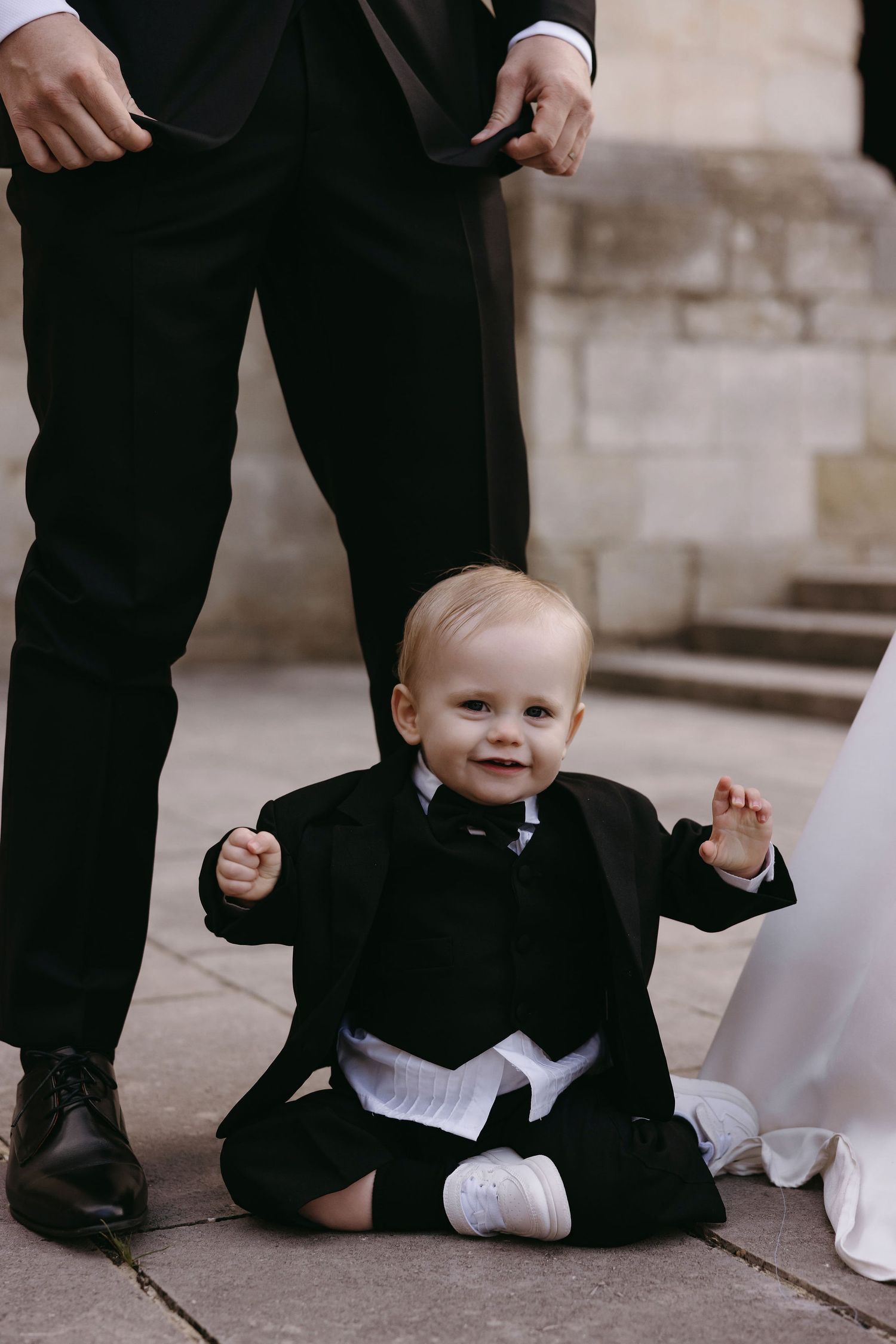 Adorable baby in formal black tuxedo sits smiling on stone ground during wedding ceremony.