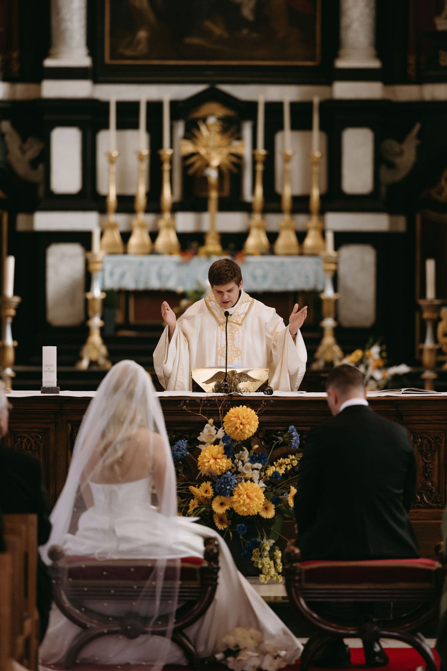 A priest conducts a wedding ceremony at an ornate church altar with golden candelabras.