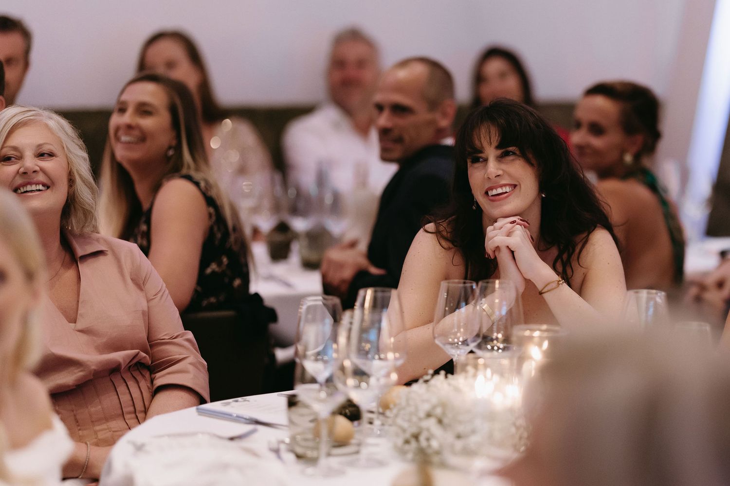 Wedding reception guests smile and celebrate at decorated dining tables with glowing candles.