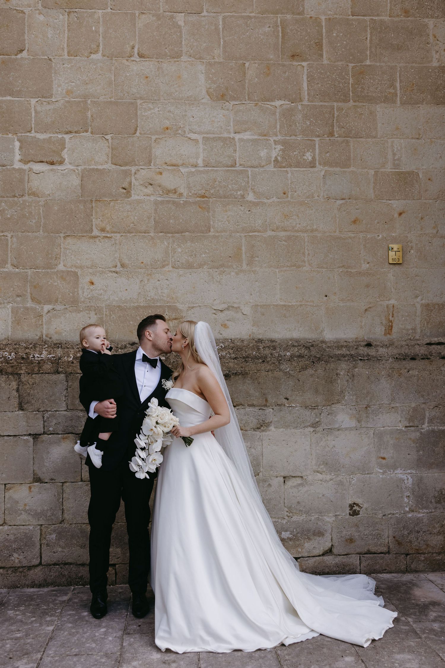Wedding portrait against a stone wall featuring elegant attire and a classic bouquet.