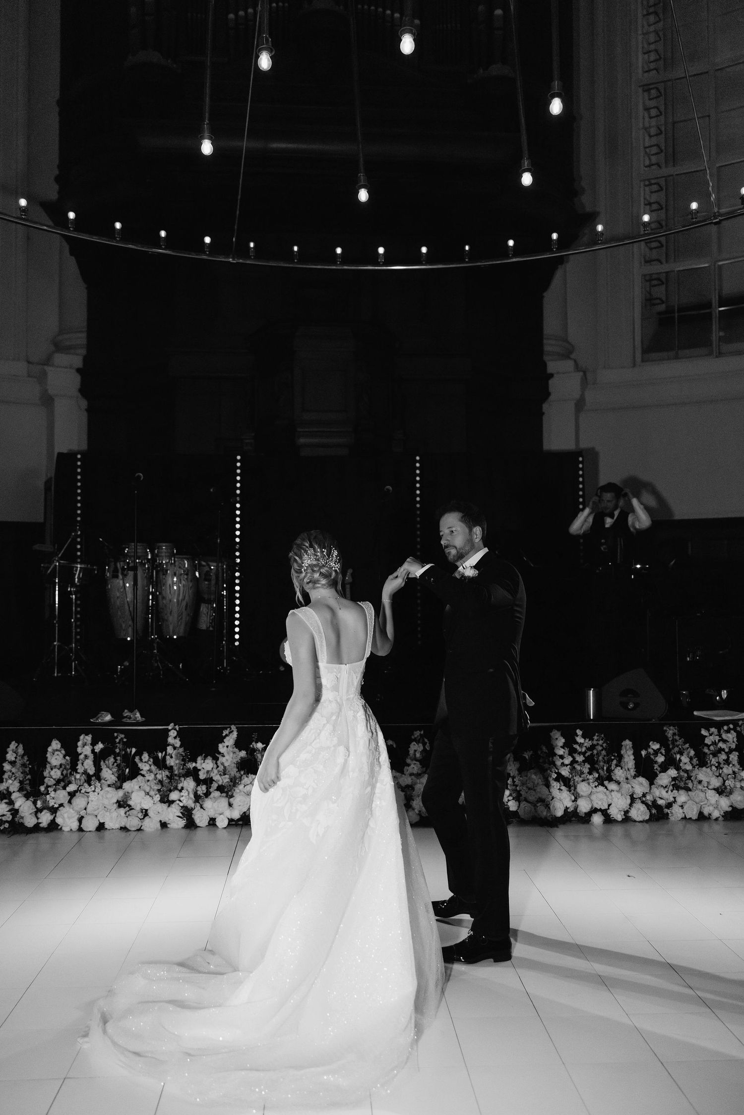 A black and white photo of a couple's first dance at their wedding reception under string lights.