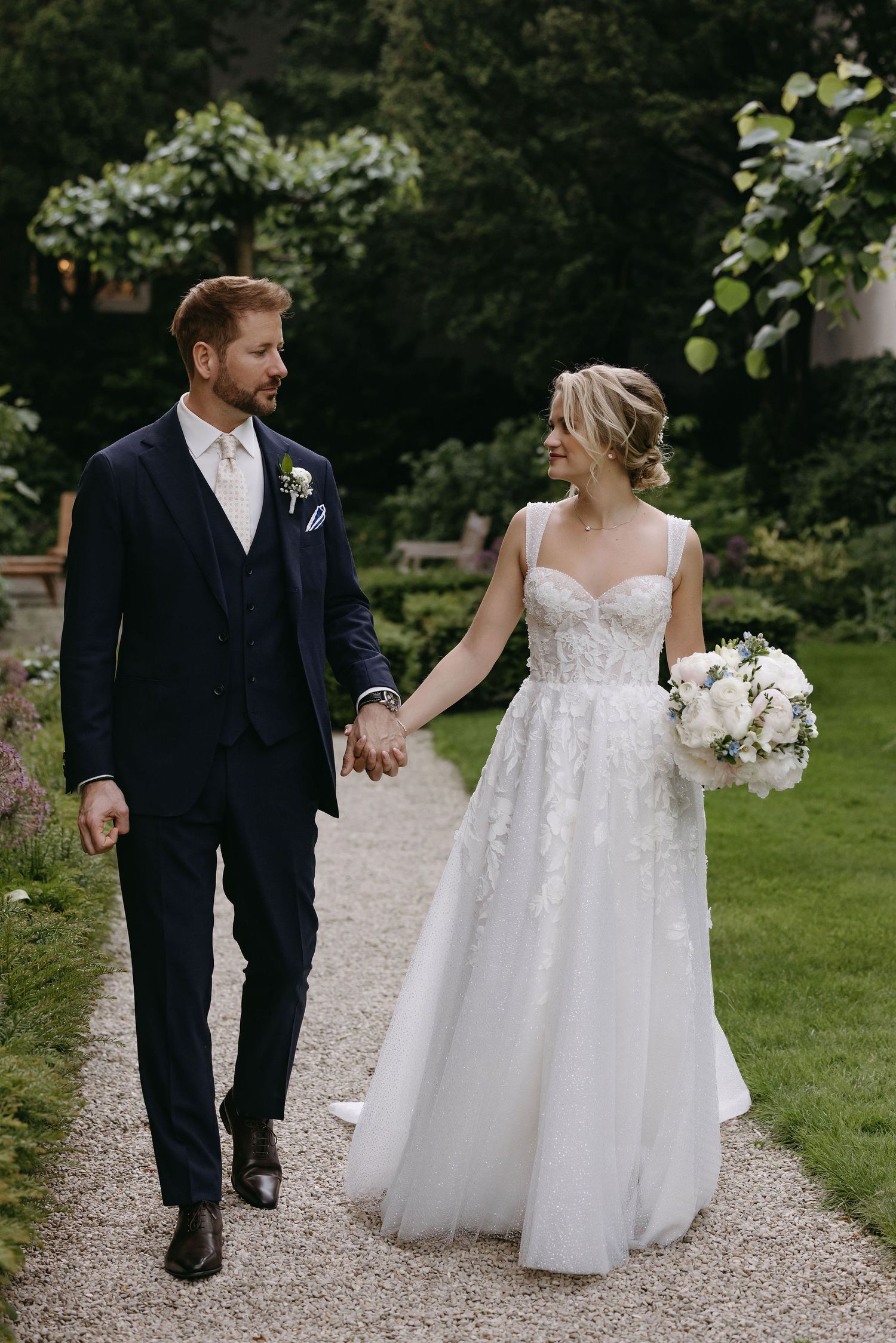 A couple strolls together on a gravel garden path holding white flower bouquets.