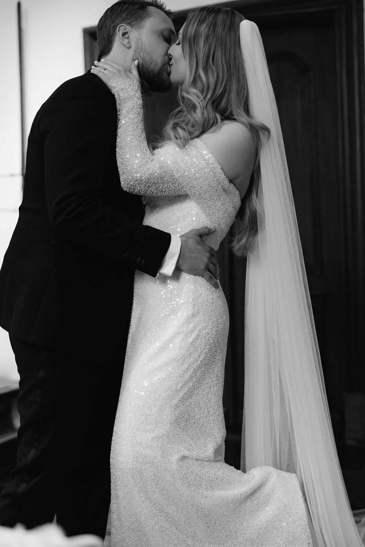 Intimate black and white portrait of newlyweds embracing, highlighting the bride's detailed gown.