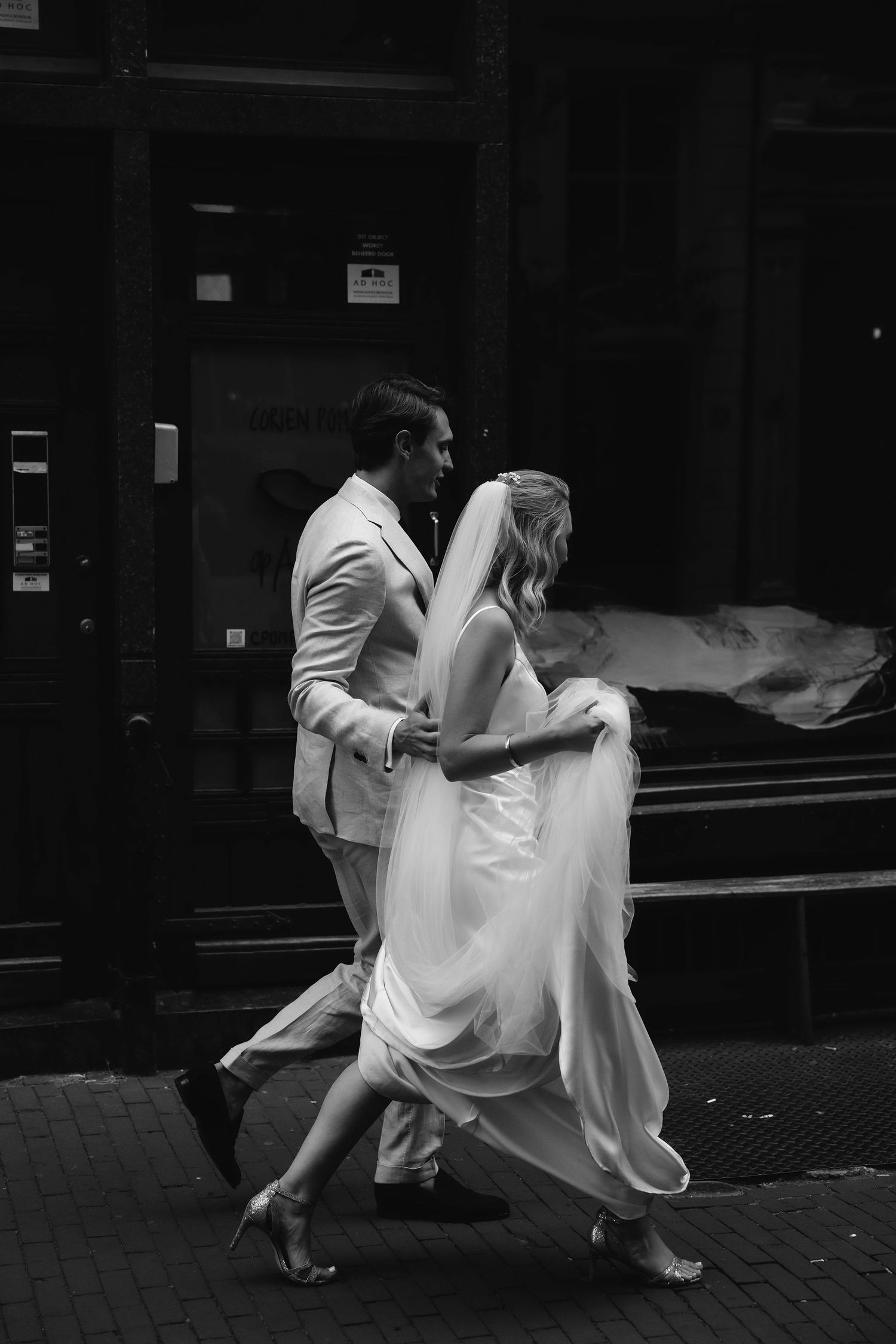 A couple in wedding attire runs playfully down an urban street in black and white.