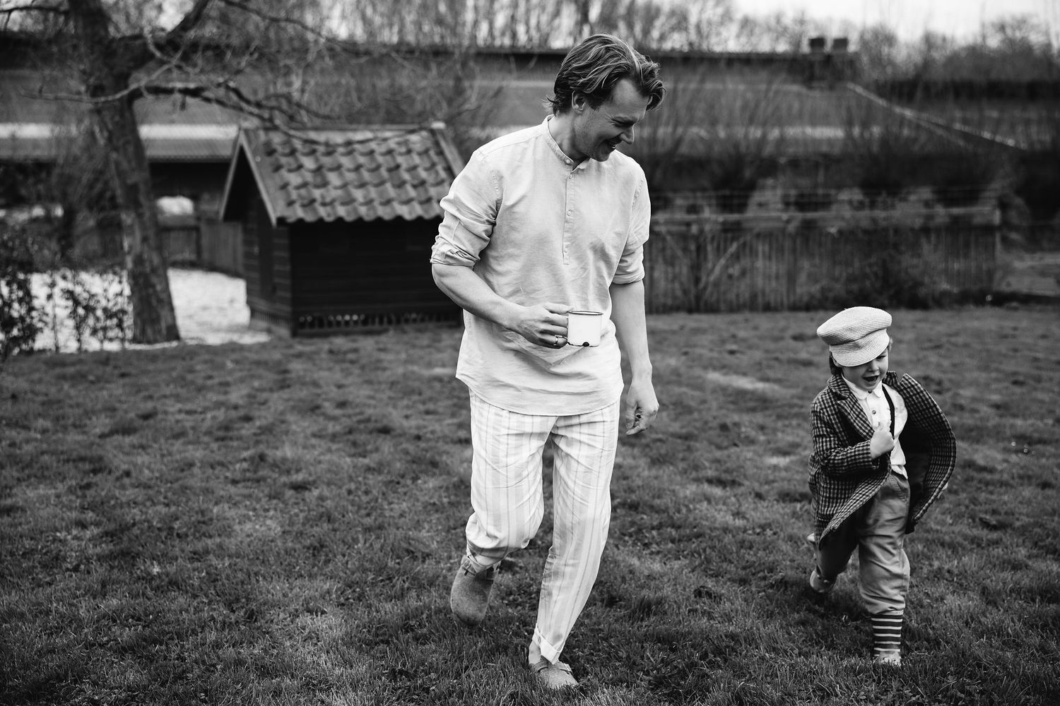 A black and white photo of two figures playing and running together on a grassy yard near small buildings.