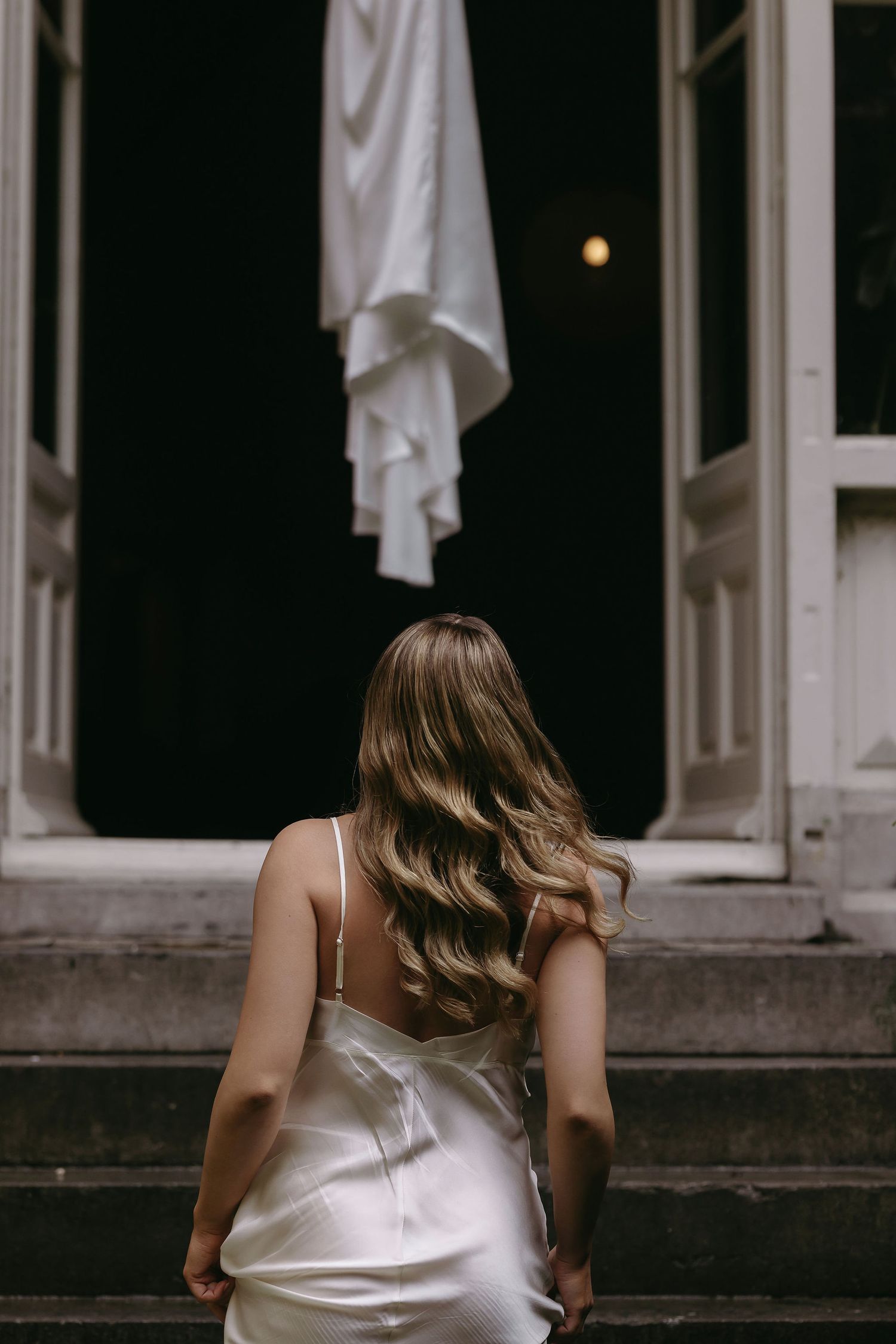 Bride in silk dress walking up stone steps towards hanging wedding gown.