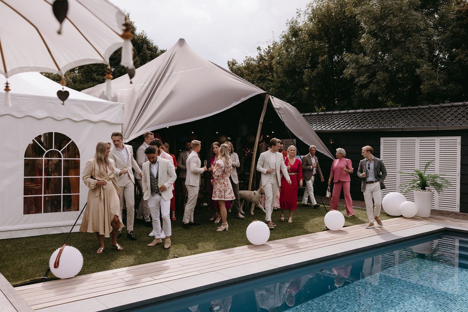 Wedding guests in formal attire gather on a white deck by a swimming pool under an elegant tent structure.