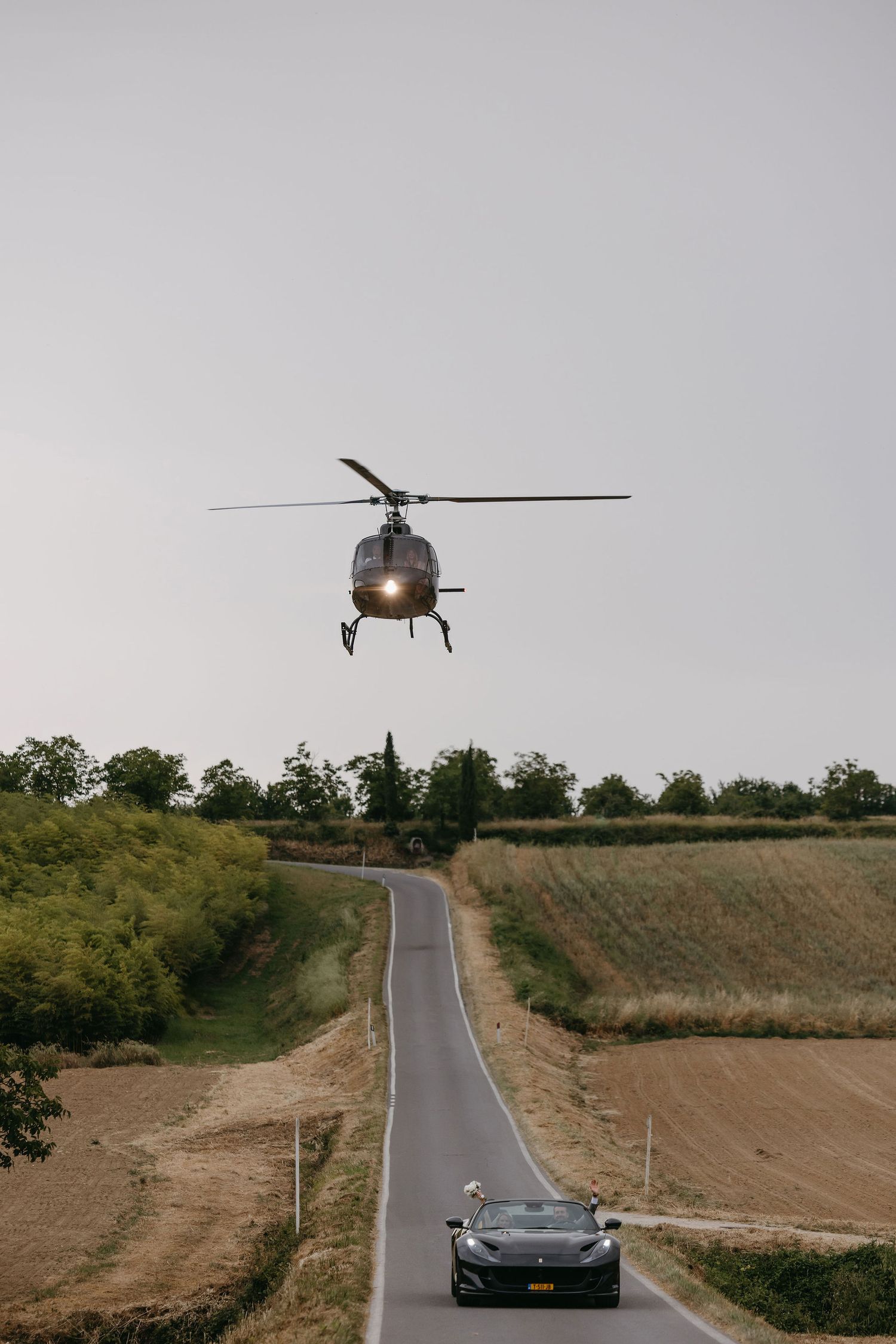 A helicopter hovers over a rural road with a sports car driving beneath it on a scenic country route.