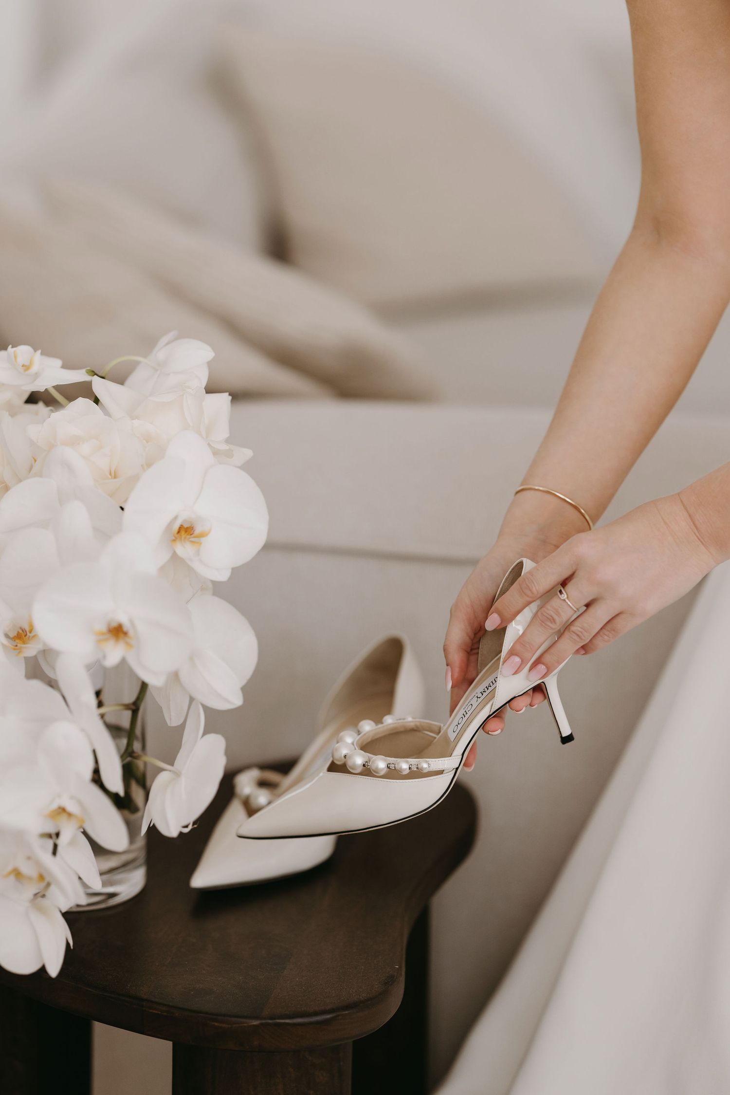 A delicate white heeled shoe sits beside an orchid arrangement as a hand with a gold bracelet reaches towards it.