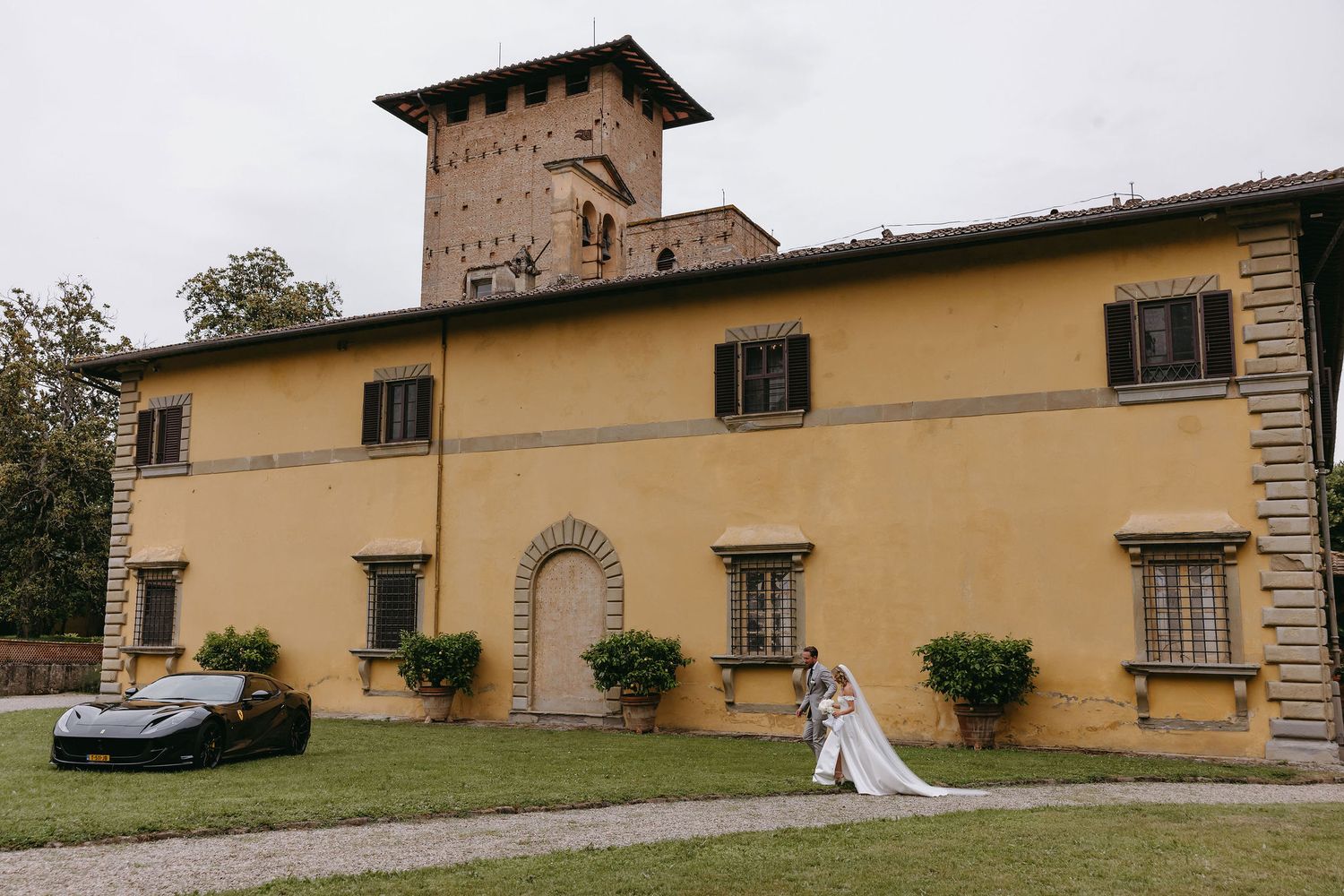 An elegant Tuscan villa with a tower and cream-colored facade serves as a romantic wedding venue with parked car.