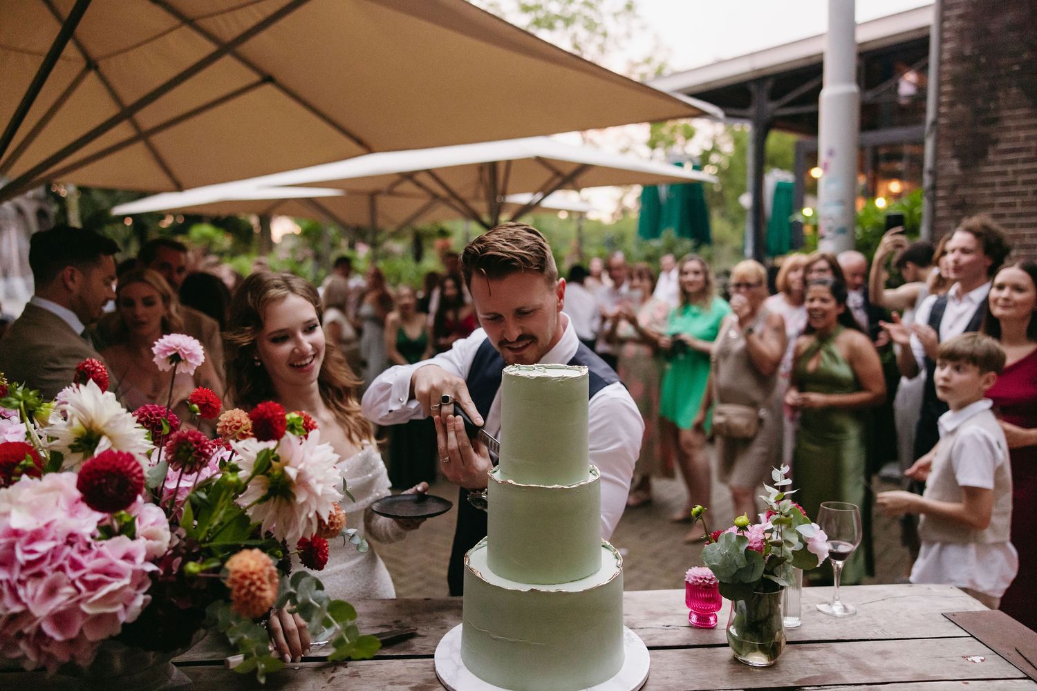 Modern white wedding cake decorated with fresh flowers at an outdoor reception venue.
