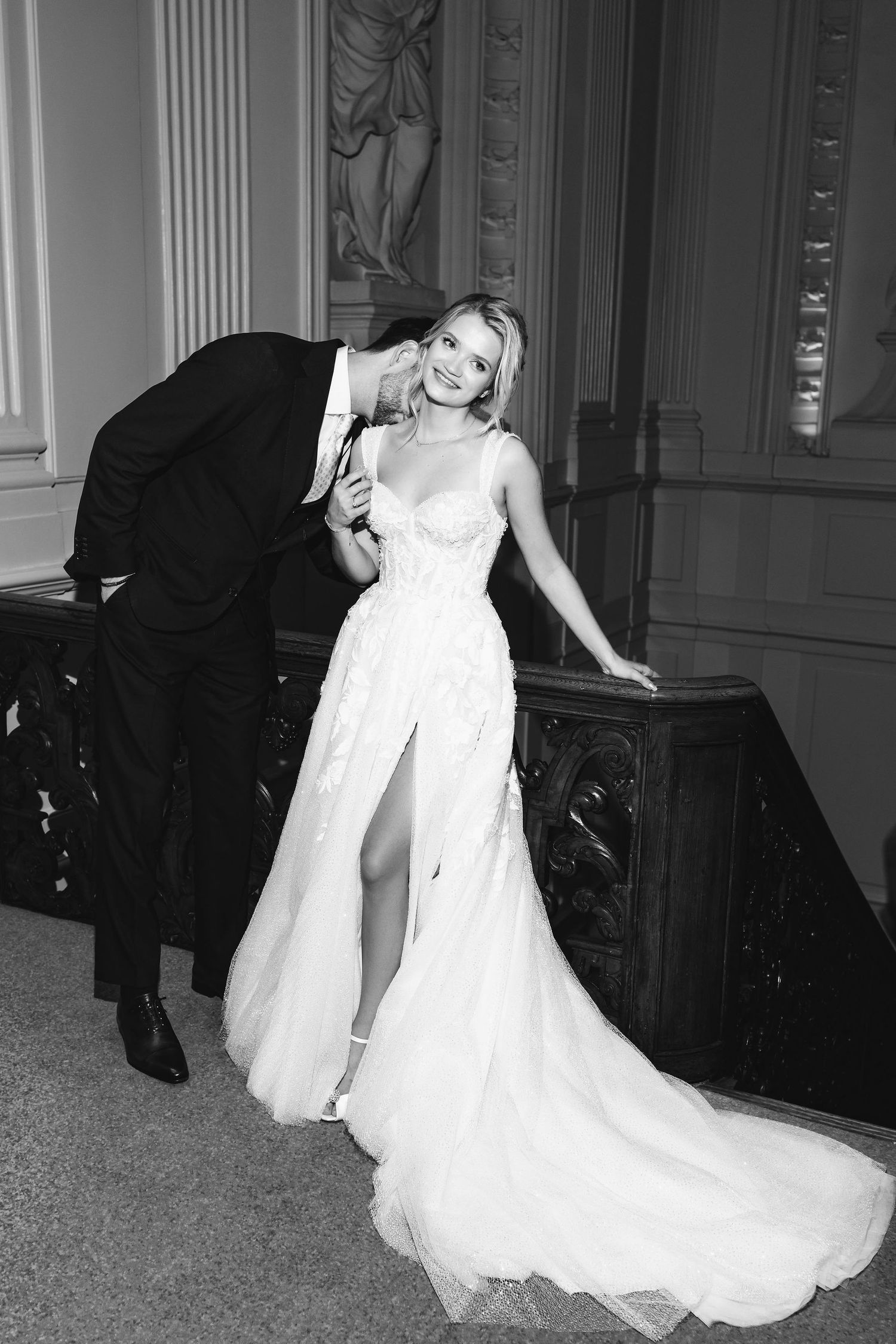 Artistic black and white portrait of couple in formal attire posed by ornate staircase.