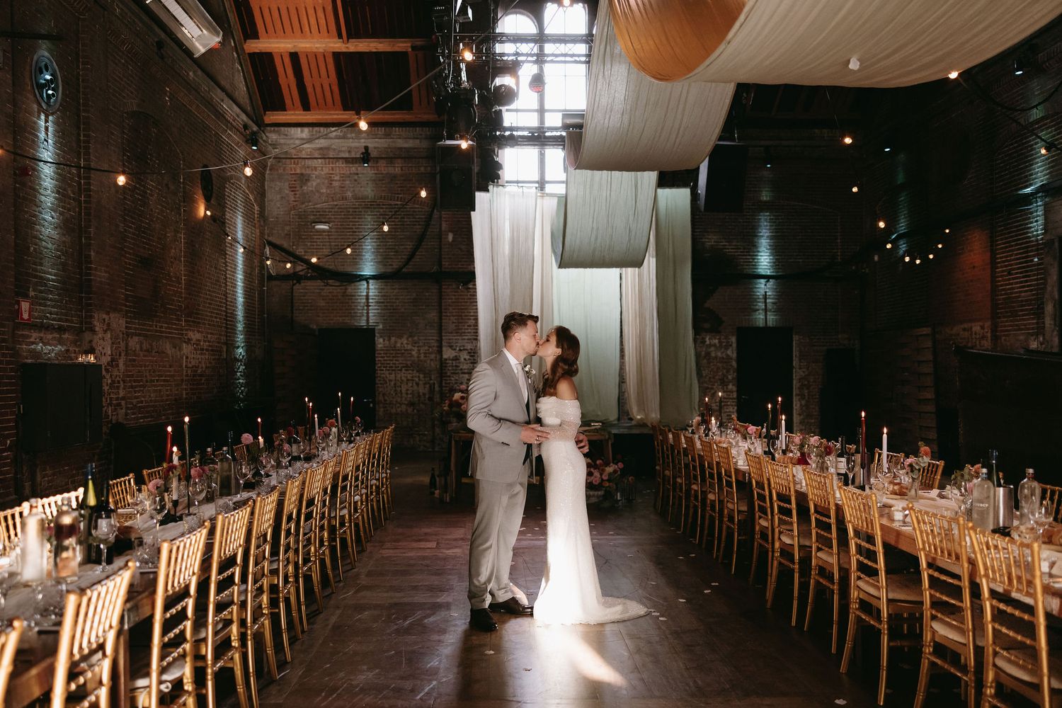 Dramatic wedding portrait of couple standing in long reception hall with wooden tables.