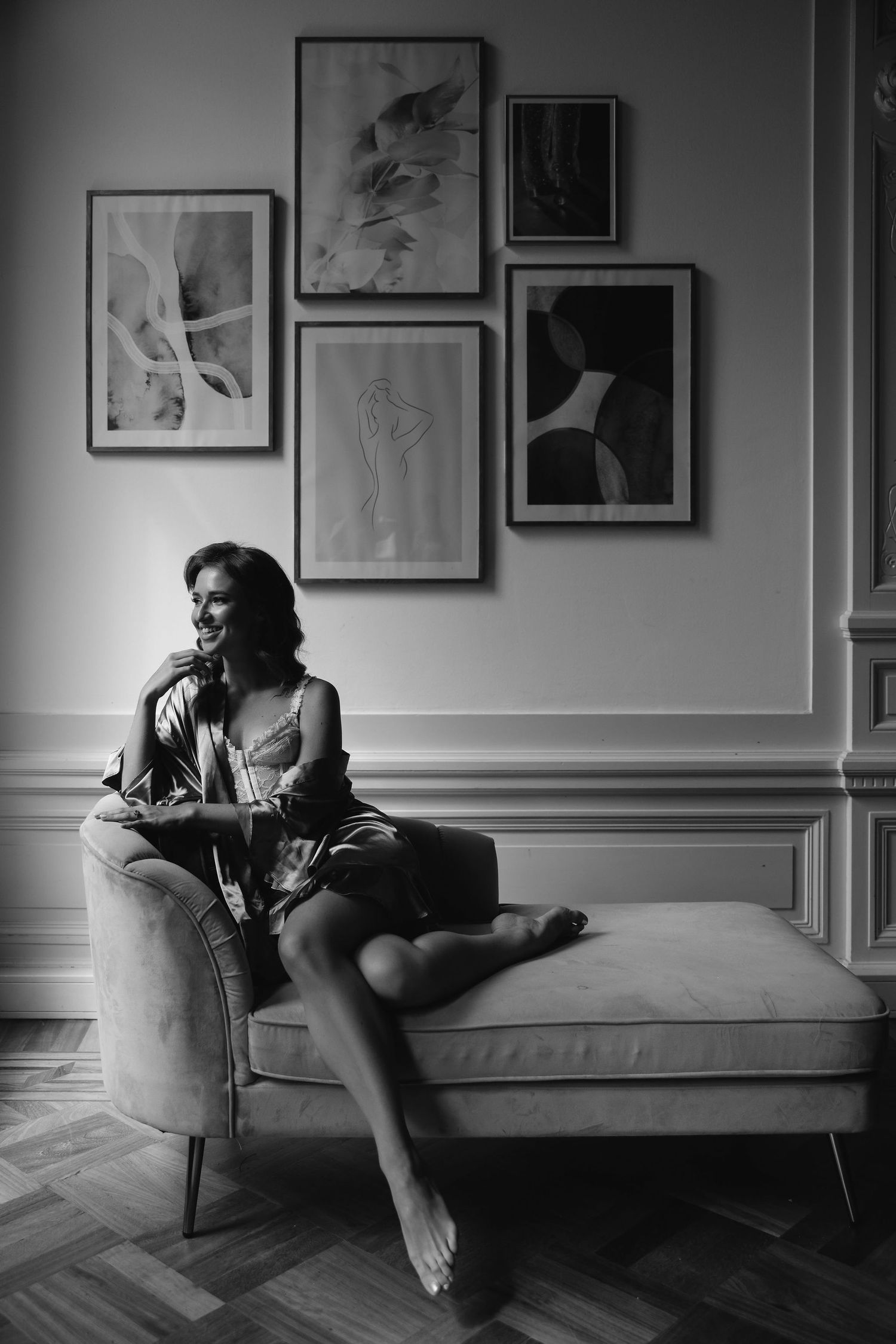 Black and white portrait of someone lounging on a chaise with artistic gallery wall behind.
