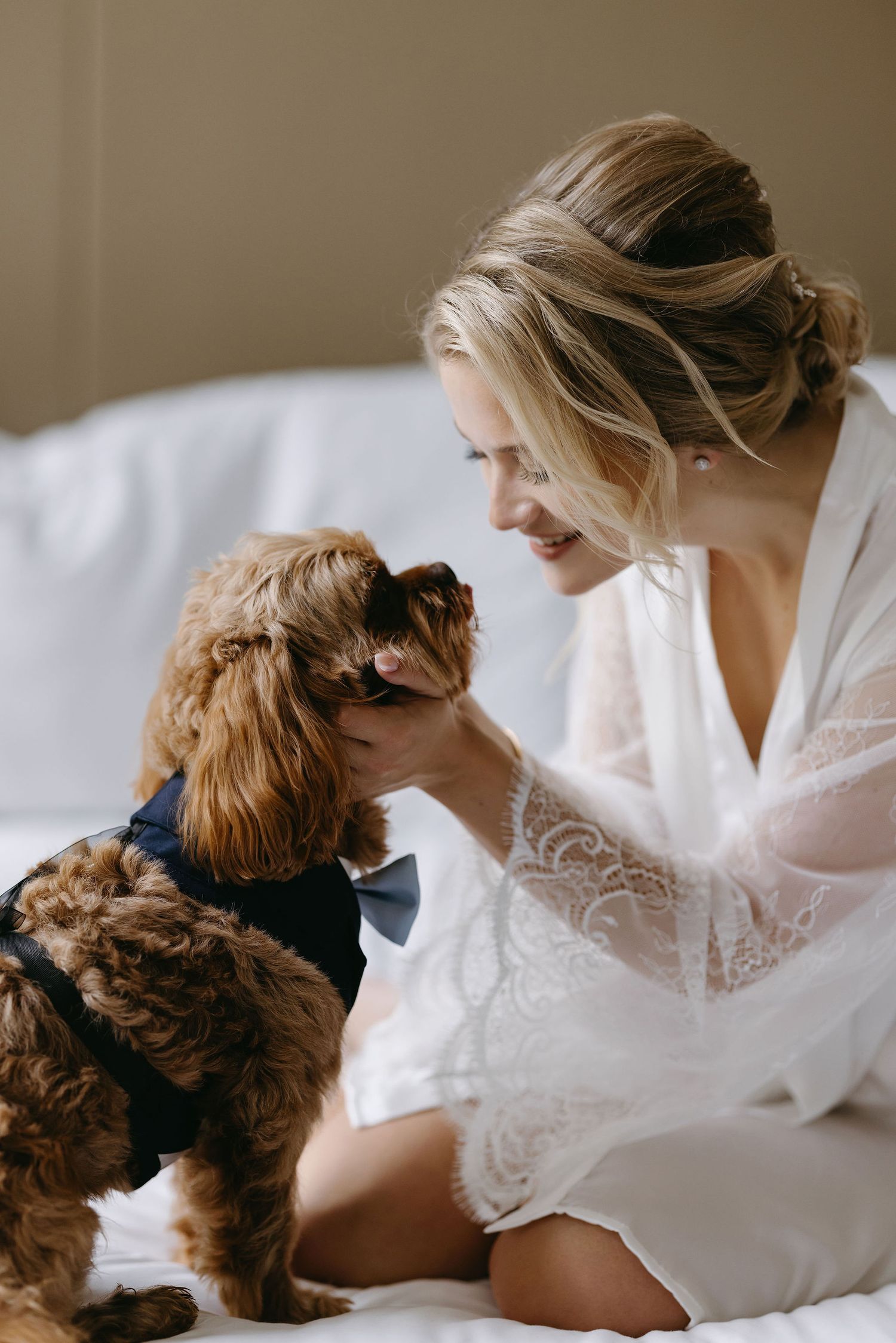 Sweet moment between person in white lace robe and brown furry dog on white bedding.
