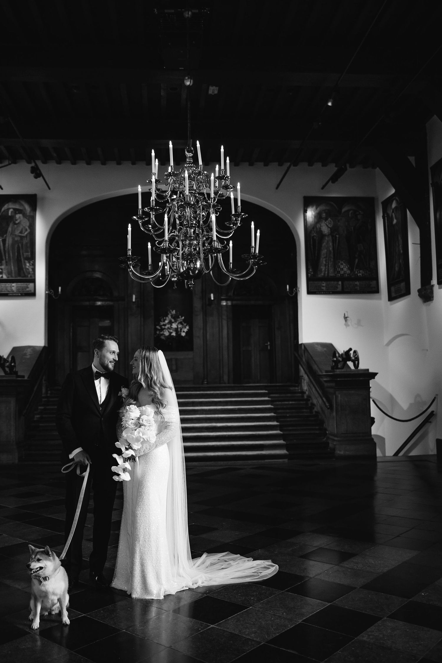 A dramatic black and white wedding photo under an ornate chandelier with a dog present.